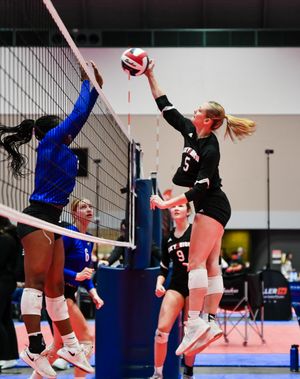 Two volleyball players compete at the net during an intense point with one attempting a block in black and blue uniforms.