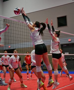 Volleyball players leap high at the net to block during an indoor match on a red court.