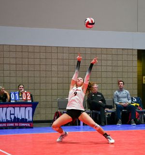Indoor volleyball player jumps to spike a red and white volleyball during a competitive match on a red court.