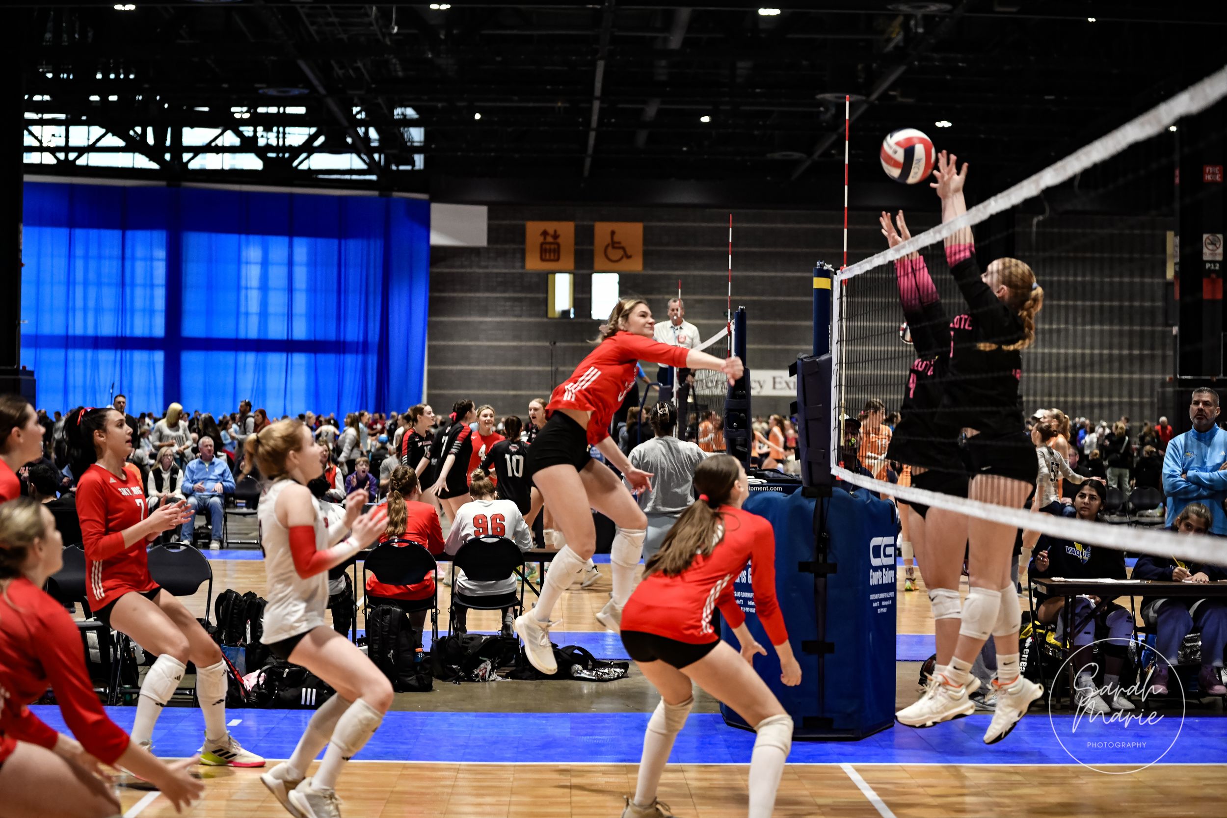 Players jump high above the net during an intense volleyball match on a blue court.