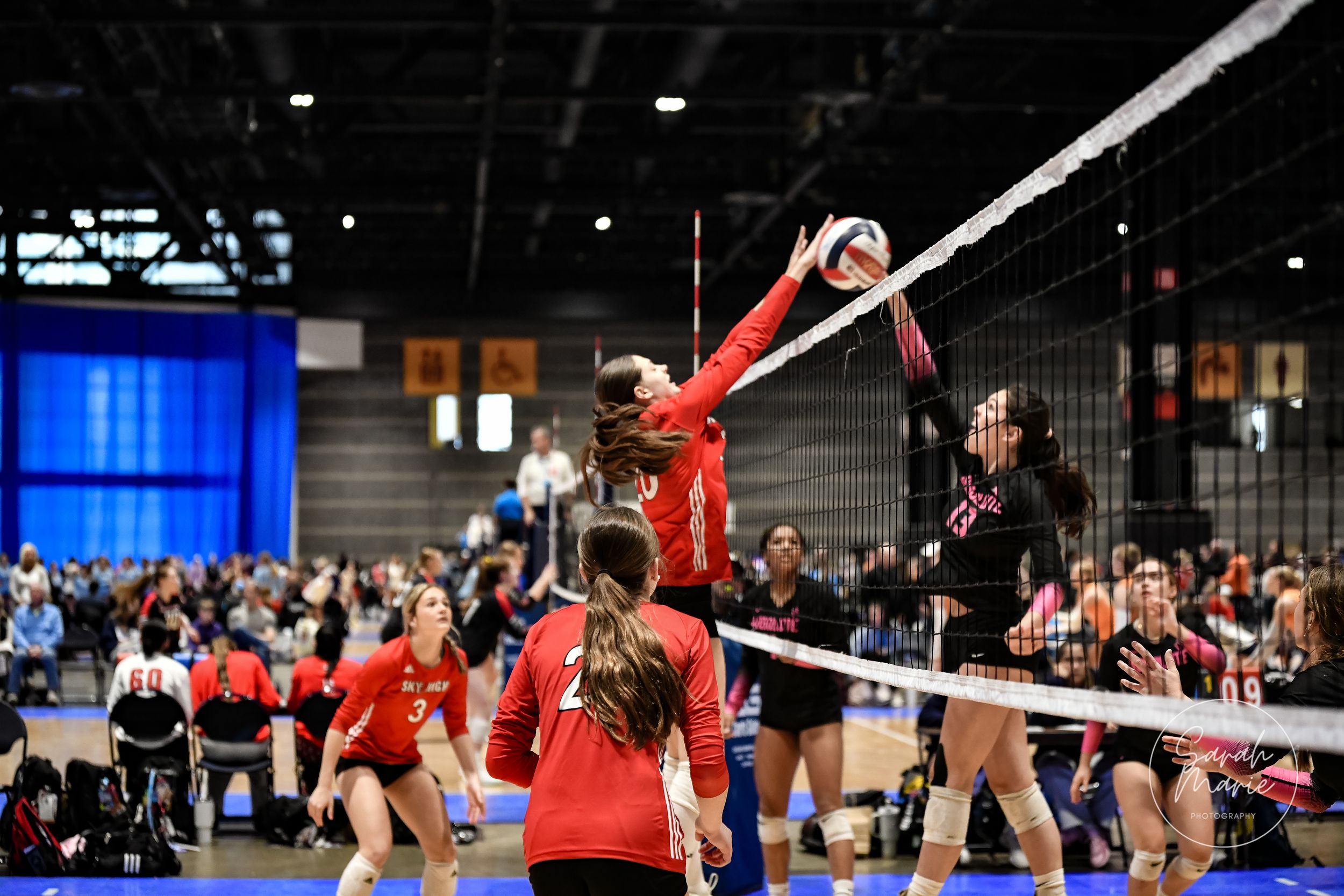 Indoor volleyball match action shot showing players jumping at the net during an intense play at a tournament.