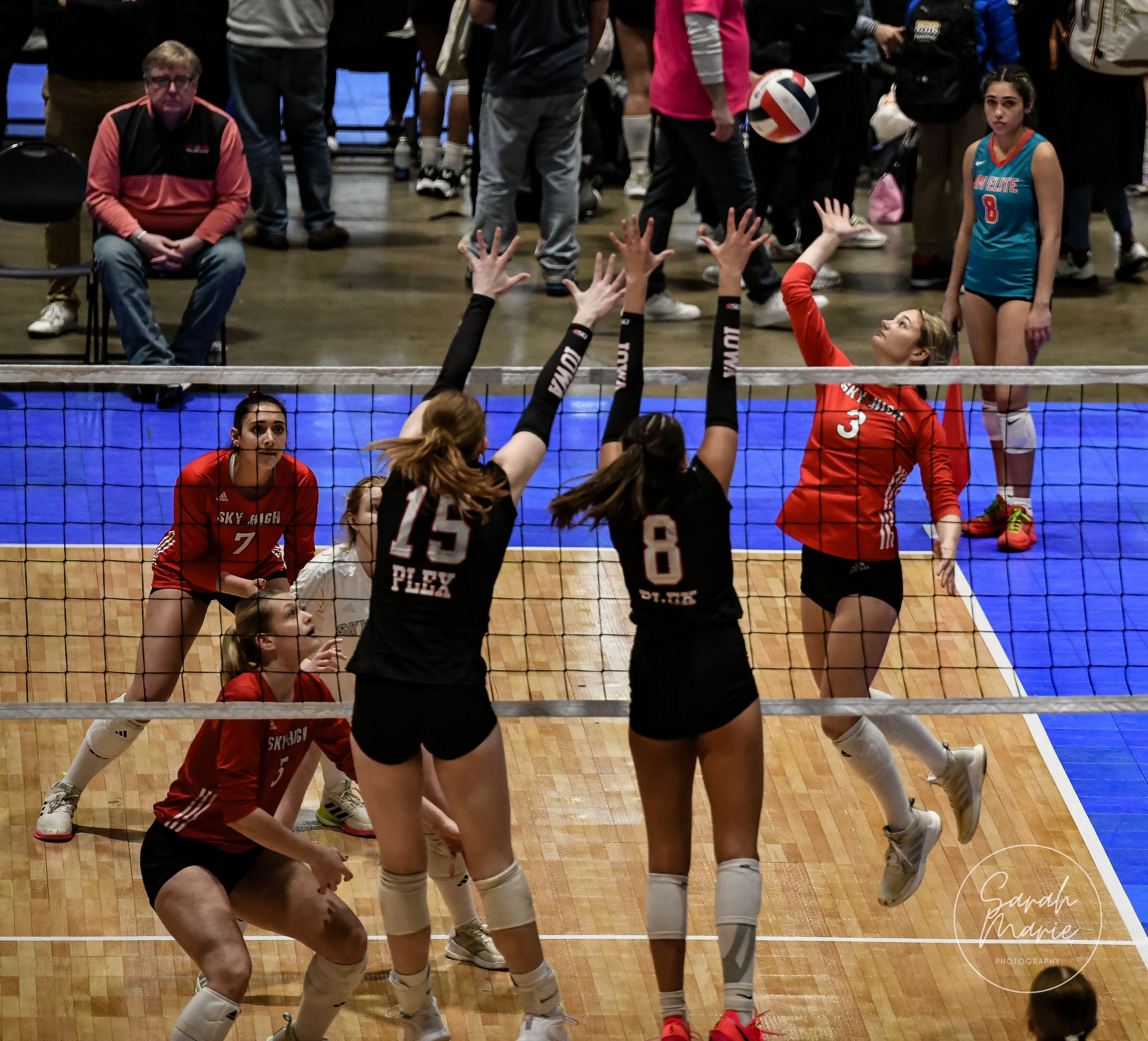 A tense volleyball match shows players at the net with one attacking while others defend on a blue-lit court.