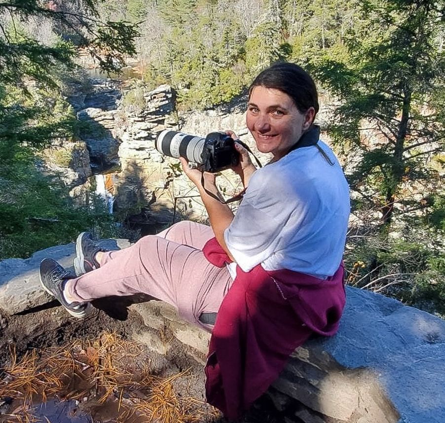 Female photographer sitting on a log with a telephoto lens camera in a rocky mountain forest.