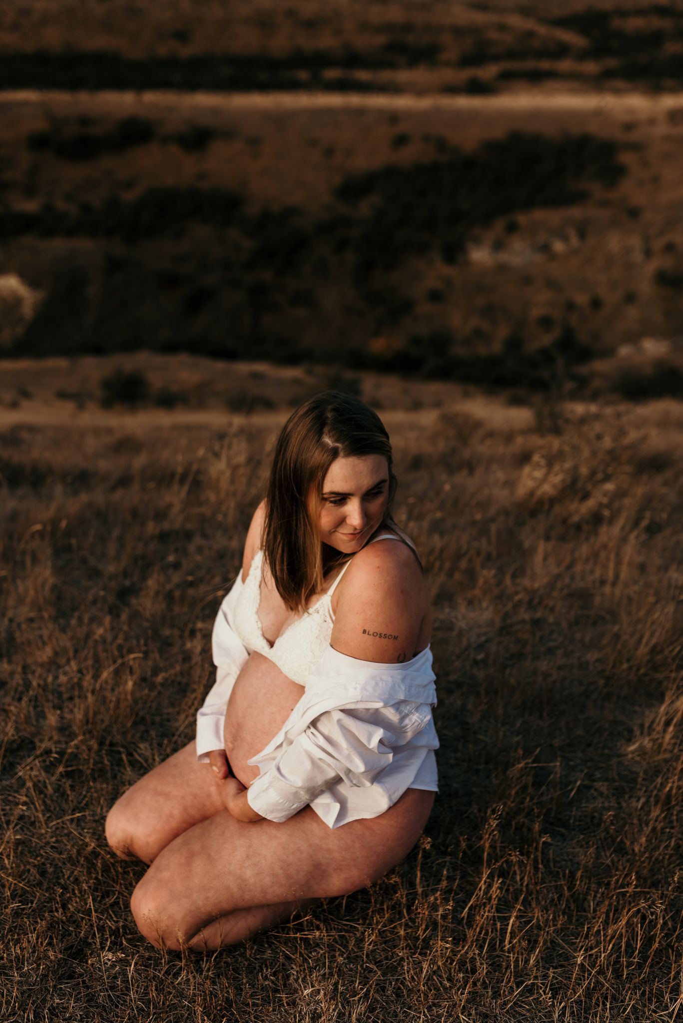 Person in white clothing sitting in dry grass field during golden hour sunset in rustic countryside setting.