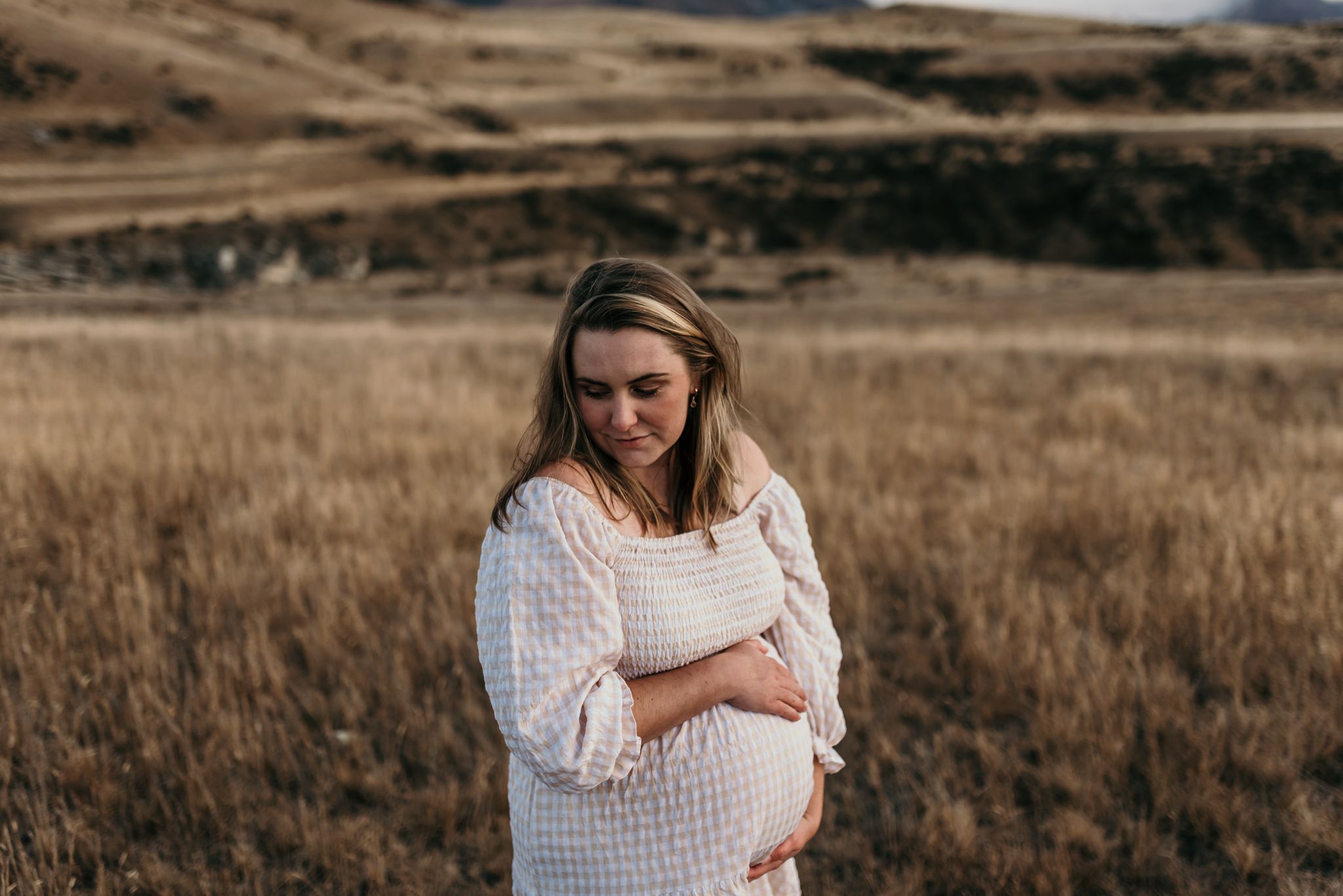 Expectant mother in white dress stands thoughtfully in dried autumn field during golden hour.