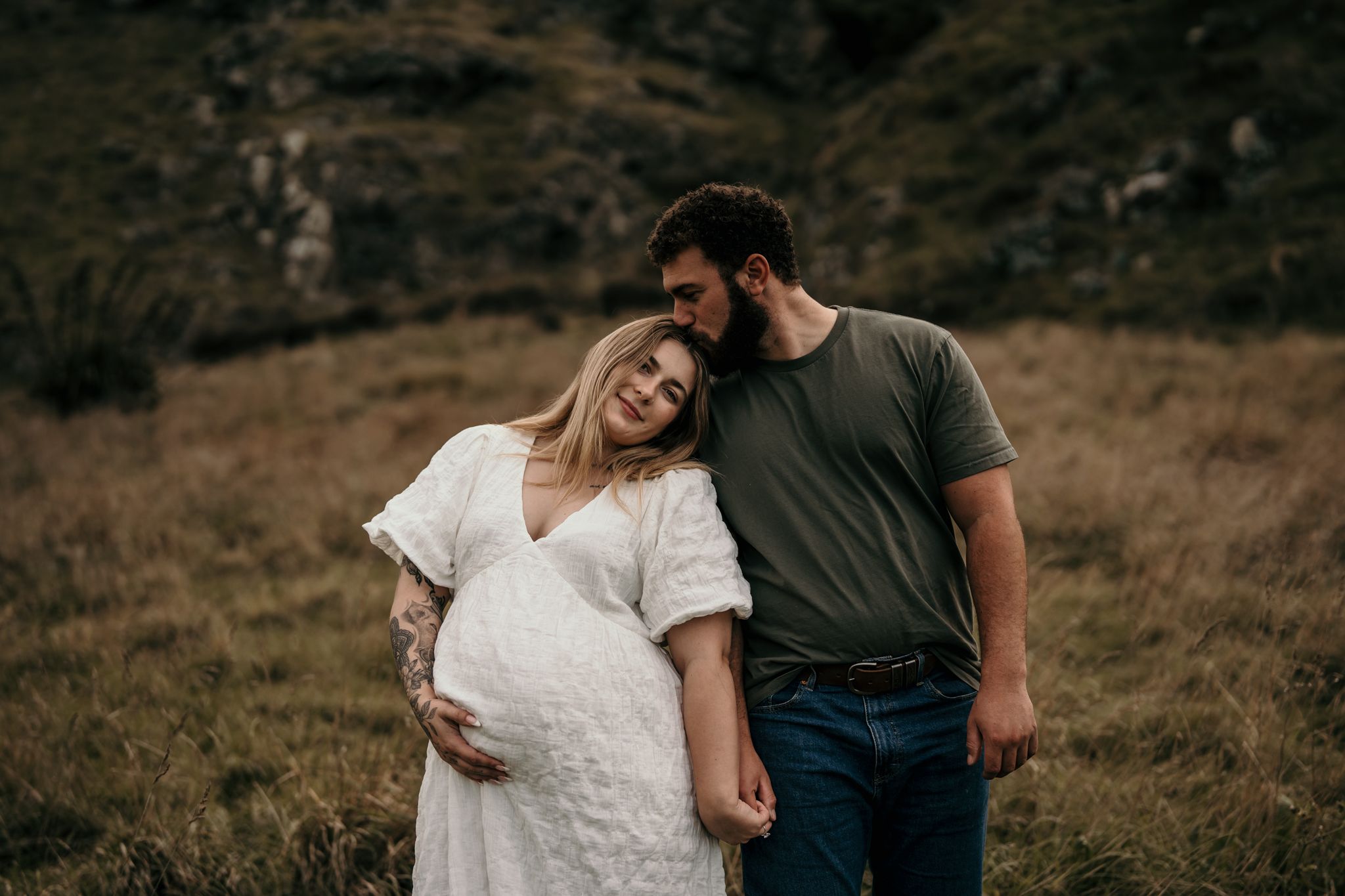 Couple shares intimate moment outdoors in mountainous field while expecting a baby.