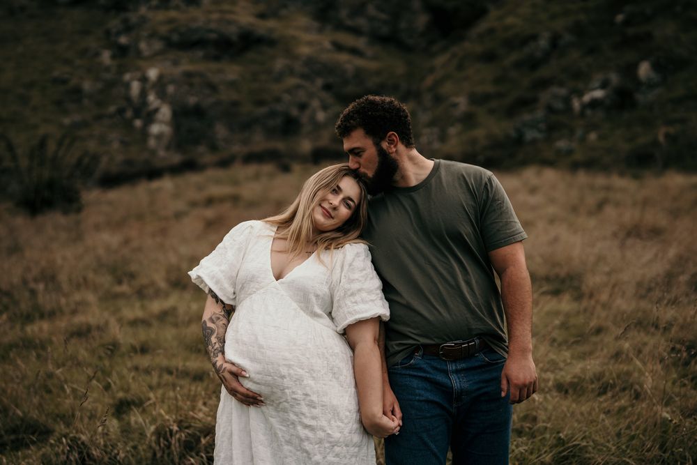 Couple shares intimate moment outdoors in mountainous field while expecting a baby.