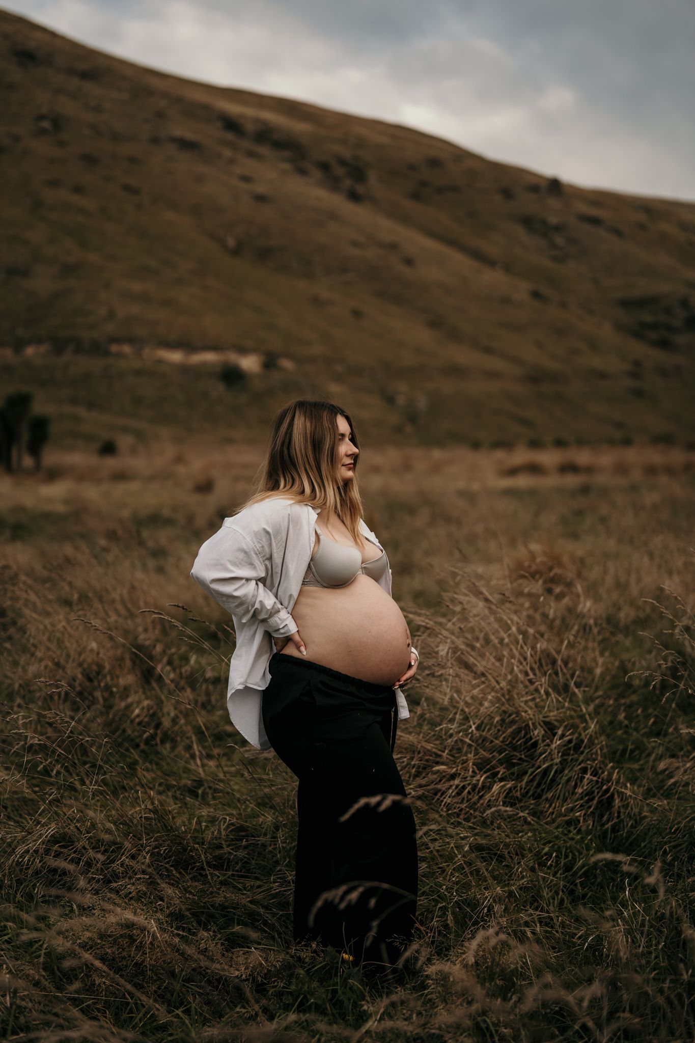 Silhouette of expecting mother in a white shirt stands against moody mountain landscape at dusk.