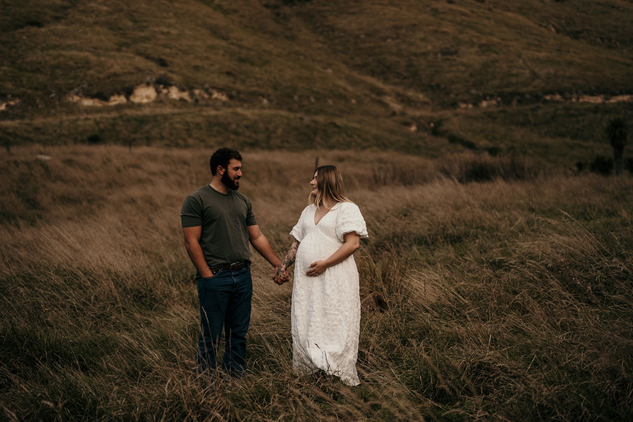 A couple stands together in a field of tall grass with rolling brown hills in the background at sunset.