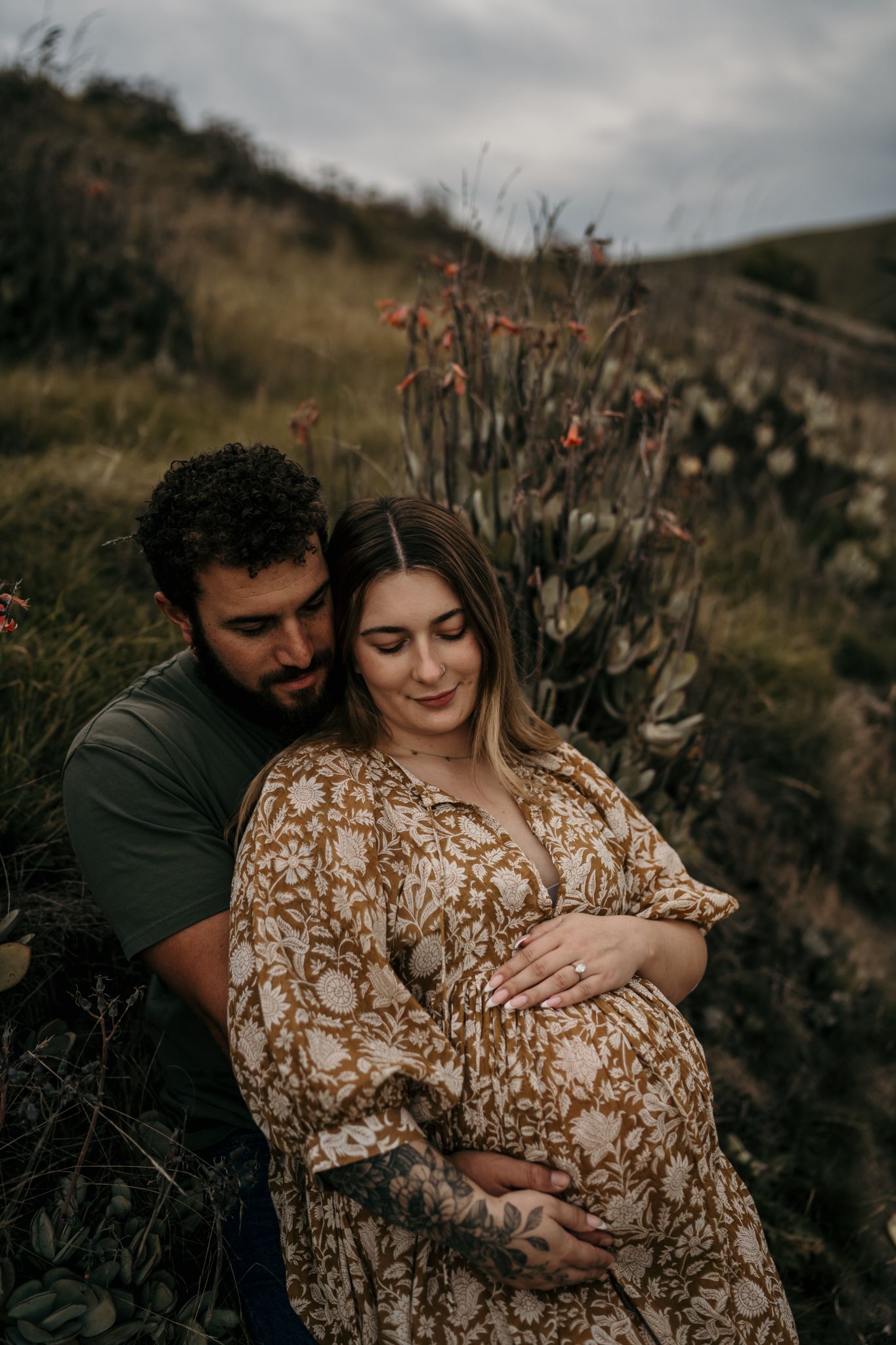 A couple embraces in a moody outdoor setting with flowers, wearing a patterned brown dress and dark shirt.