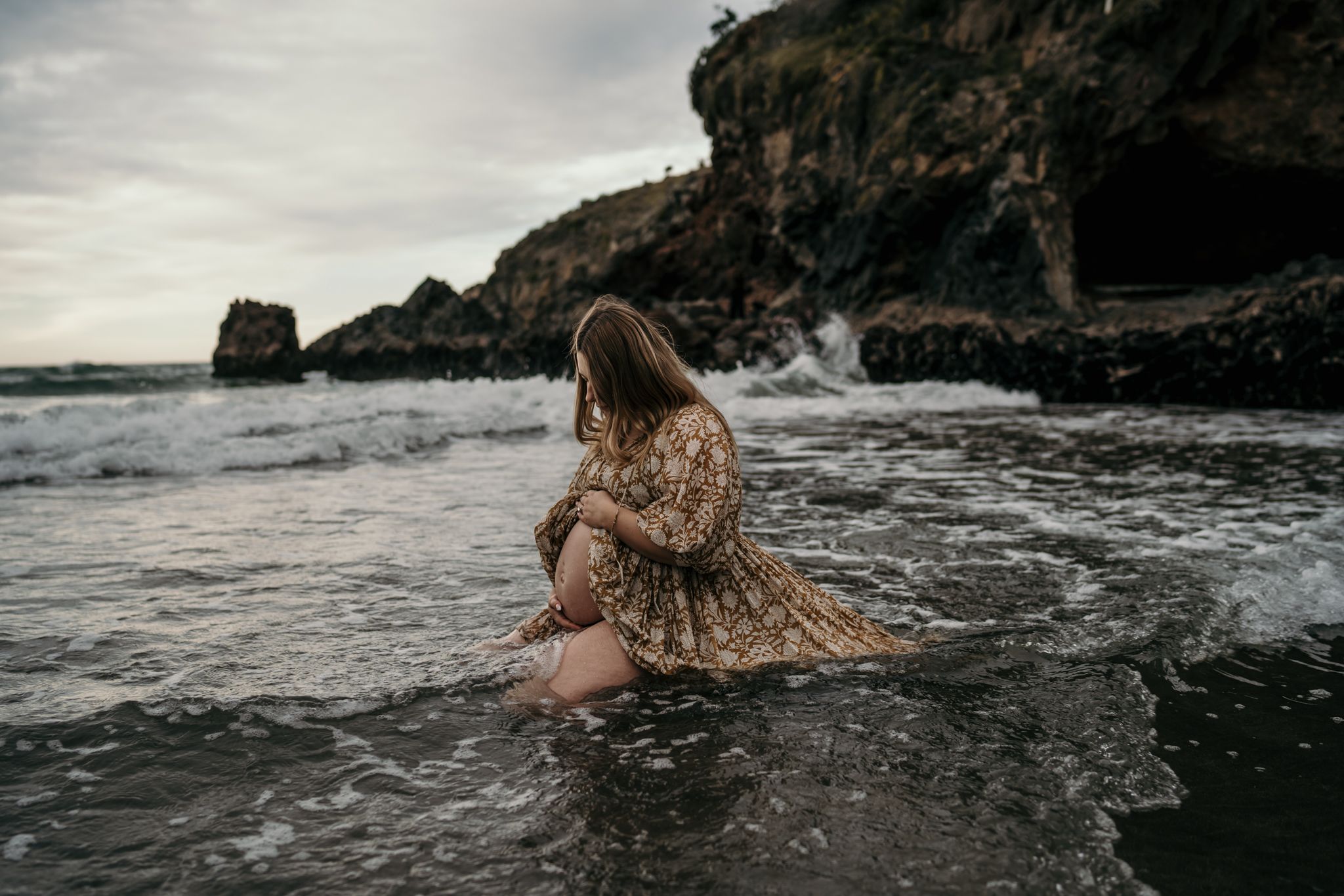 A person in a floral dress sits in shallow ocean water near rocky cliffs at sunset.