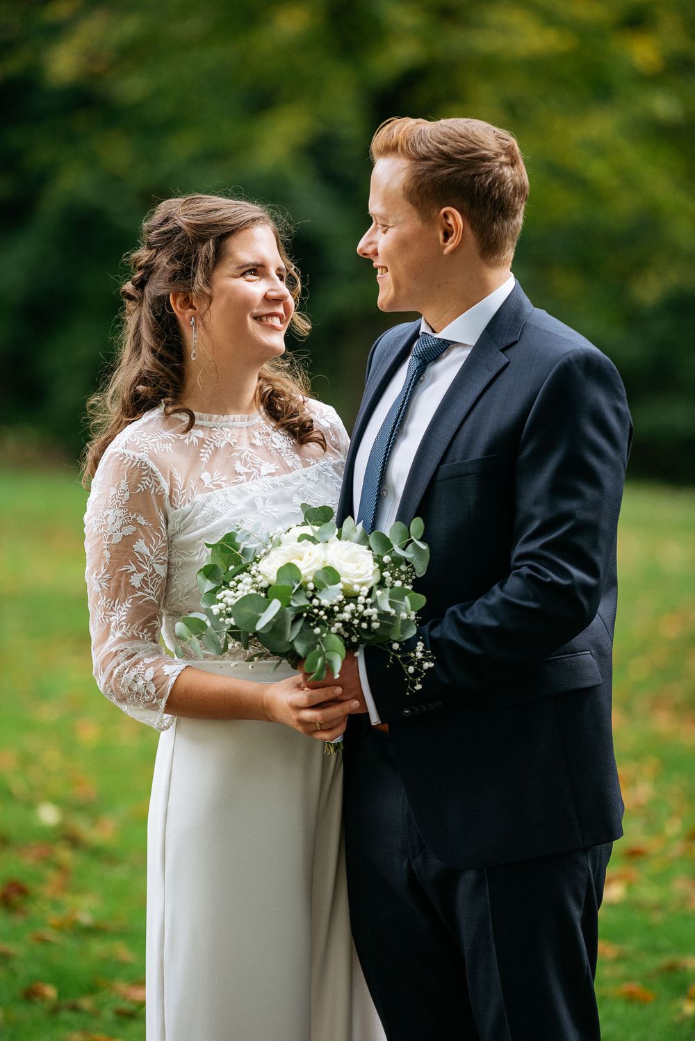 Eine Braut in einem langärmeligen Spitzenkleid teilt einen Moment mit ihrem Bräutigam und hält einen weißen Blumenstrauß im Freien.