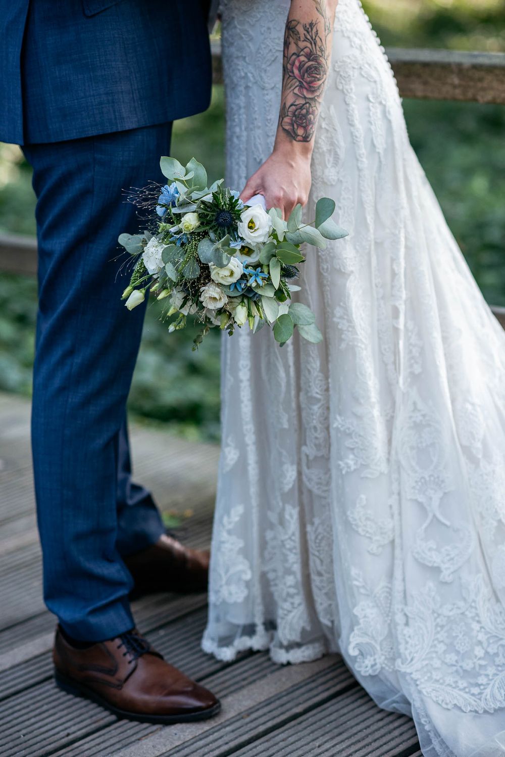 Braut im Spitzenkleid und Bräutigam im blauen Anzug halten einen blauen und weißen Blumenstrauß.