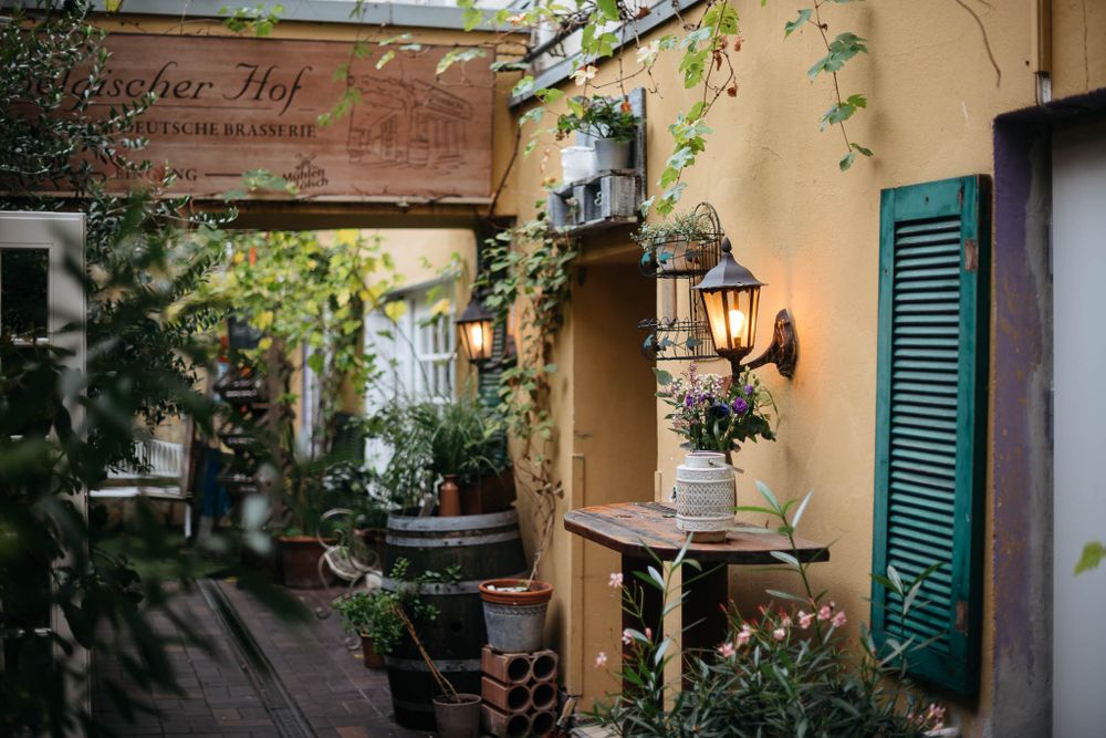 Eine charmante europäische Café-Terrasse mit grünen Fensterläden, rankenden Pflanzen und Vintage-Laternen an einer beigen Stuckwand.