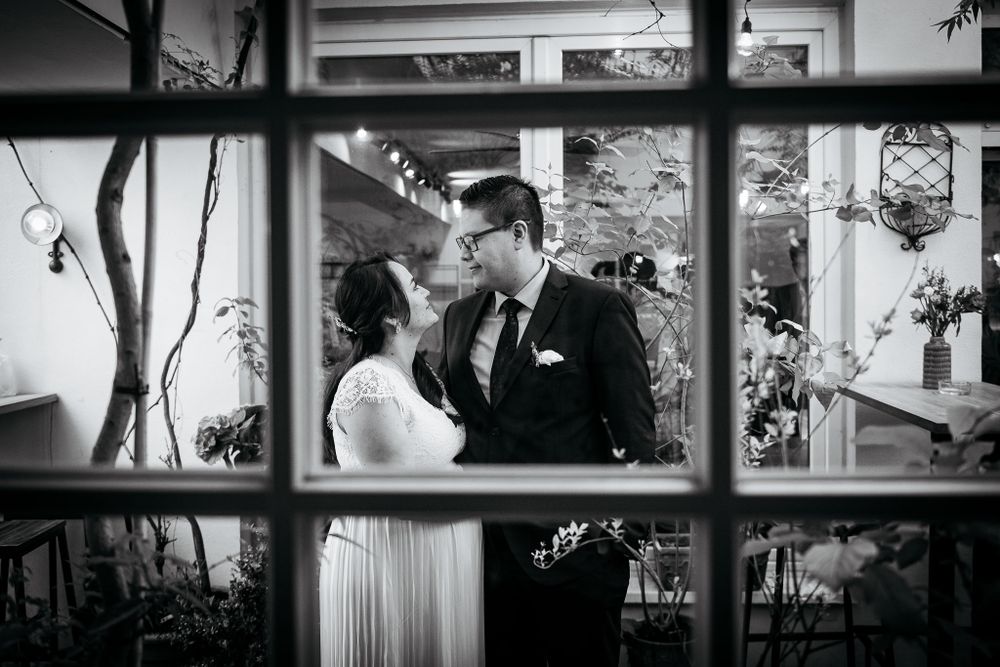 Ein romantisches Schwarz-Weiß-Foto eines Paares in formeller Kleidung, durch einen Fensterrahmen betrachtet.