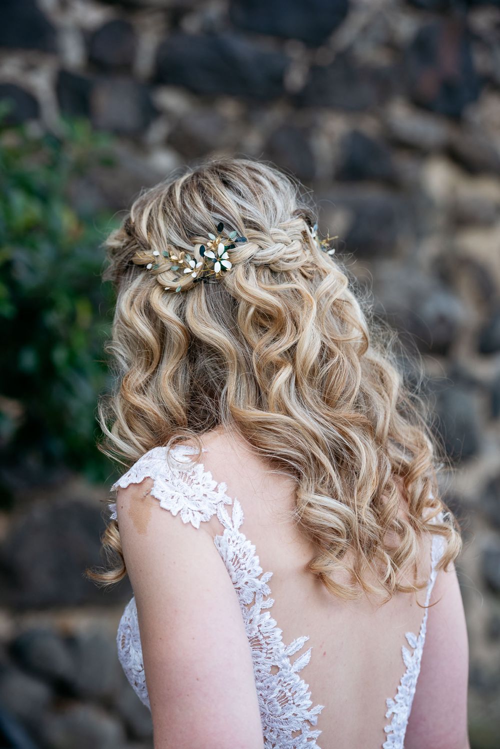 Eine Hochzeitsfrisur mit blonden Locken und einem zarten blauen Blumenaccessoire, dazu ein Spitzen-detail am Rückenteil des Kleides.