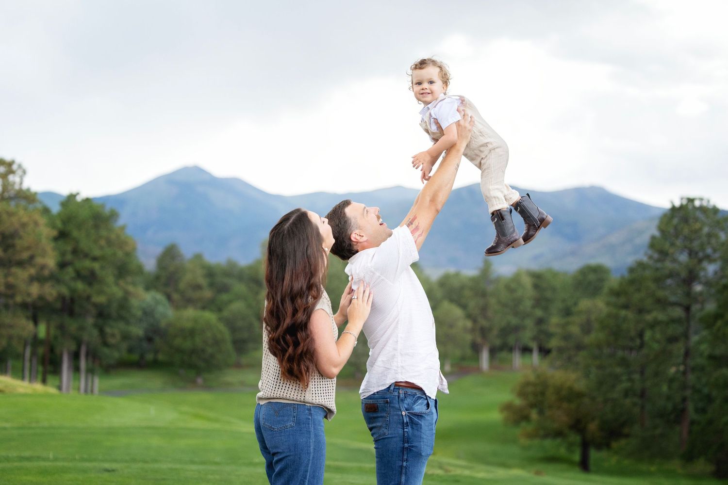 Parents lift their baby joyfully in a park at golden hour during an outdoor family photo session.