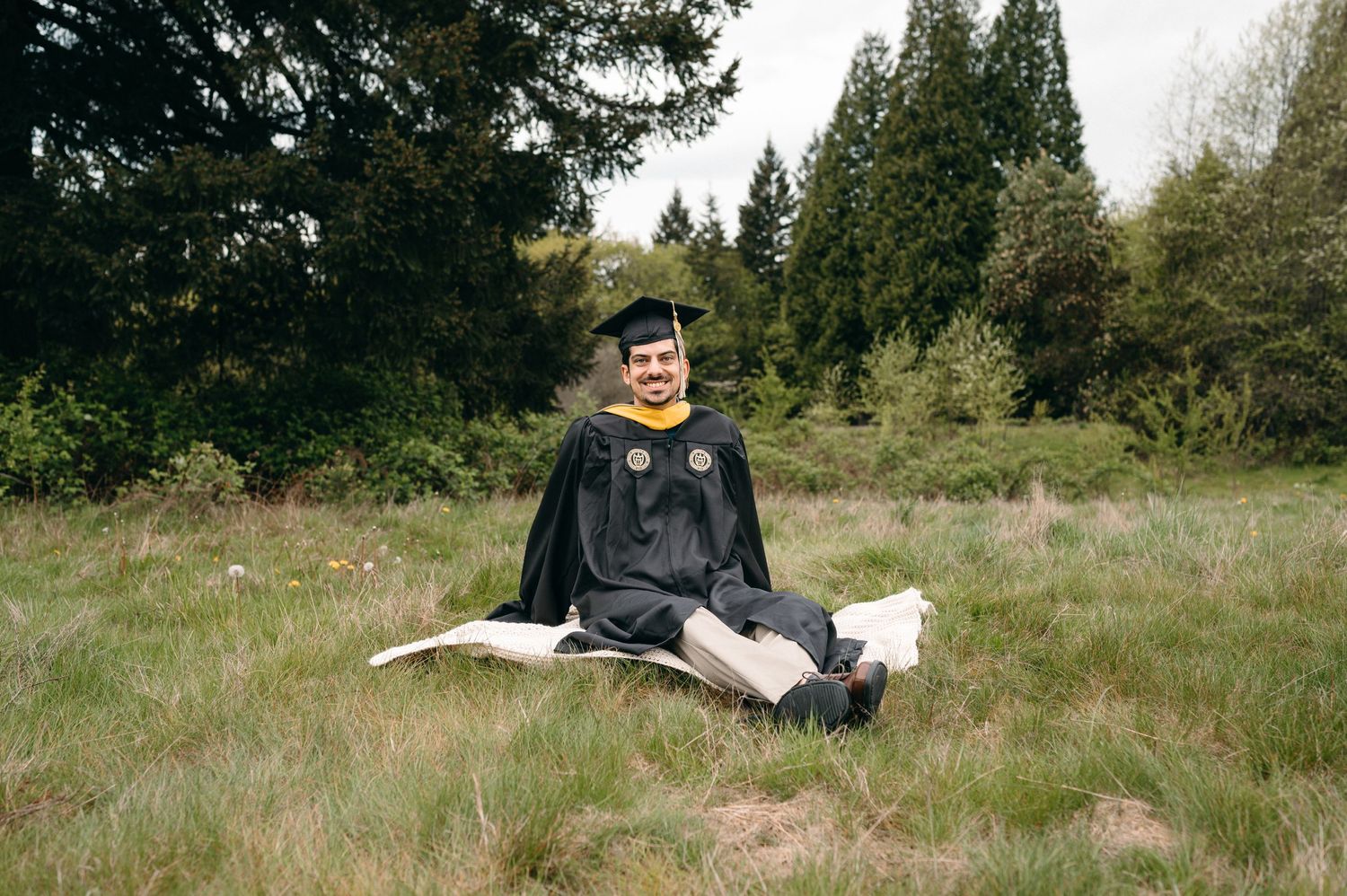 Graduate in cap and gown sitting on grass field with trees in background.