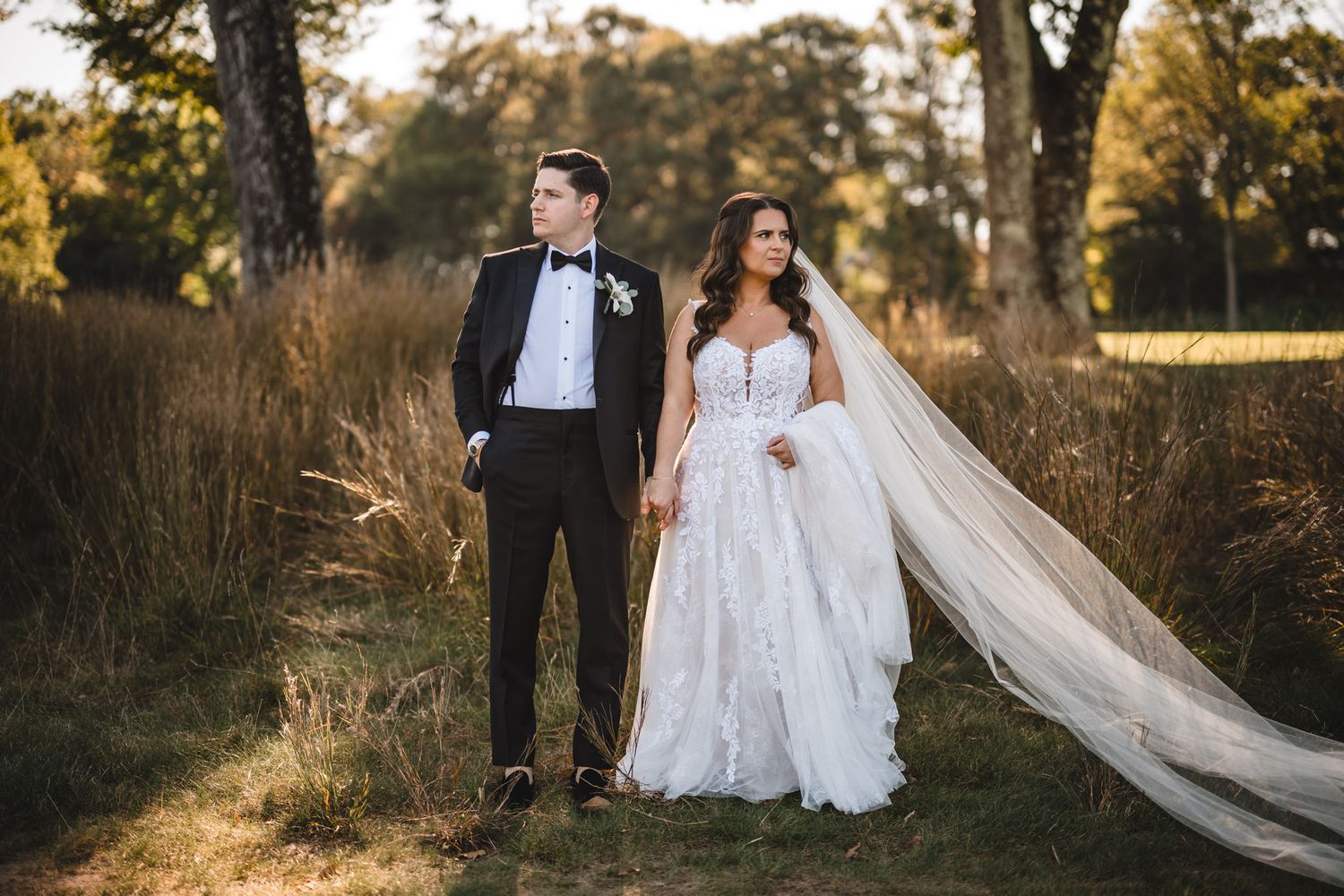 A couple in wedding attire stands together in a sunlit autumn field with a flowing veil catching the breeze.