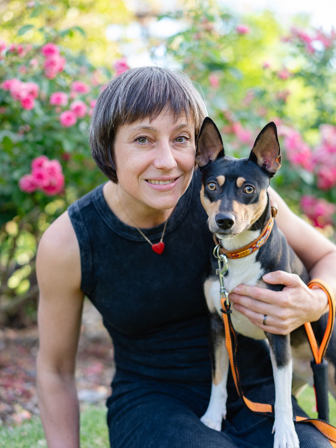 Black and tan mixed breed dog with alert ears poses with companion in front of pink flowering bush in garden.