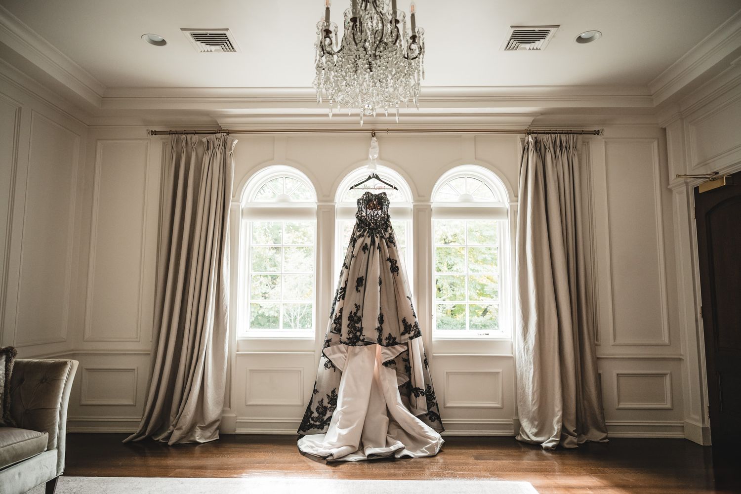 Elegant wedding dress displayed under crystal chandelier in bright room with arched windows.