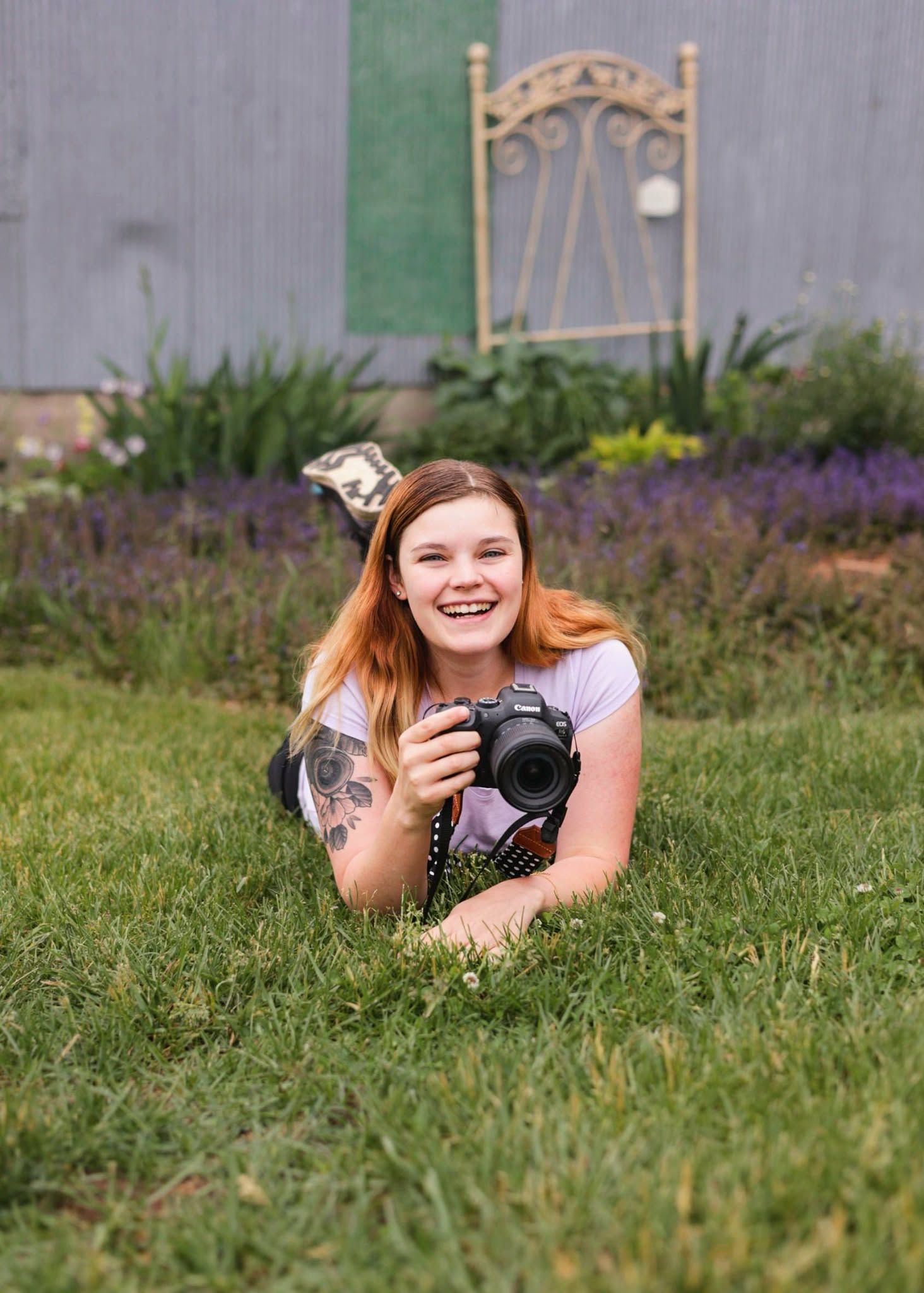 A photographer lying in purple flowers holding a camera and smiling.