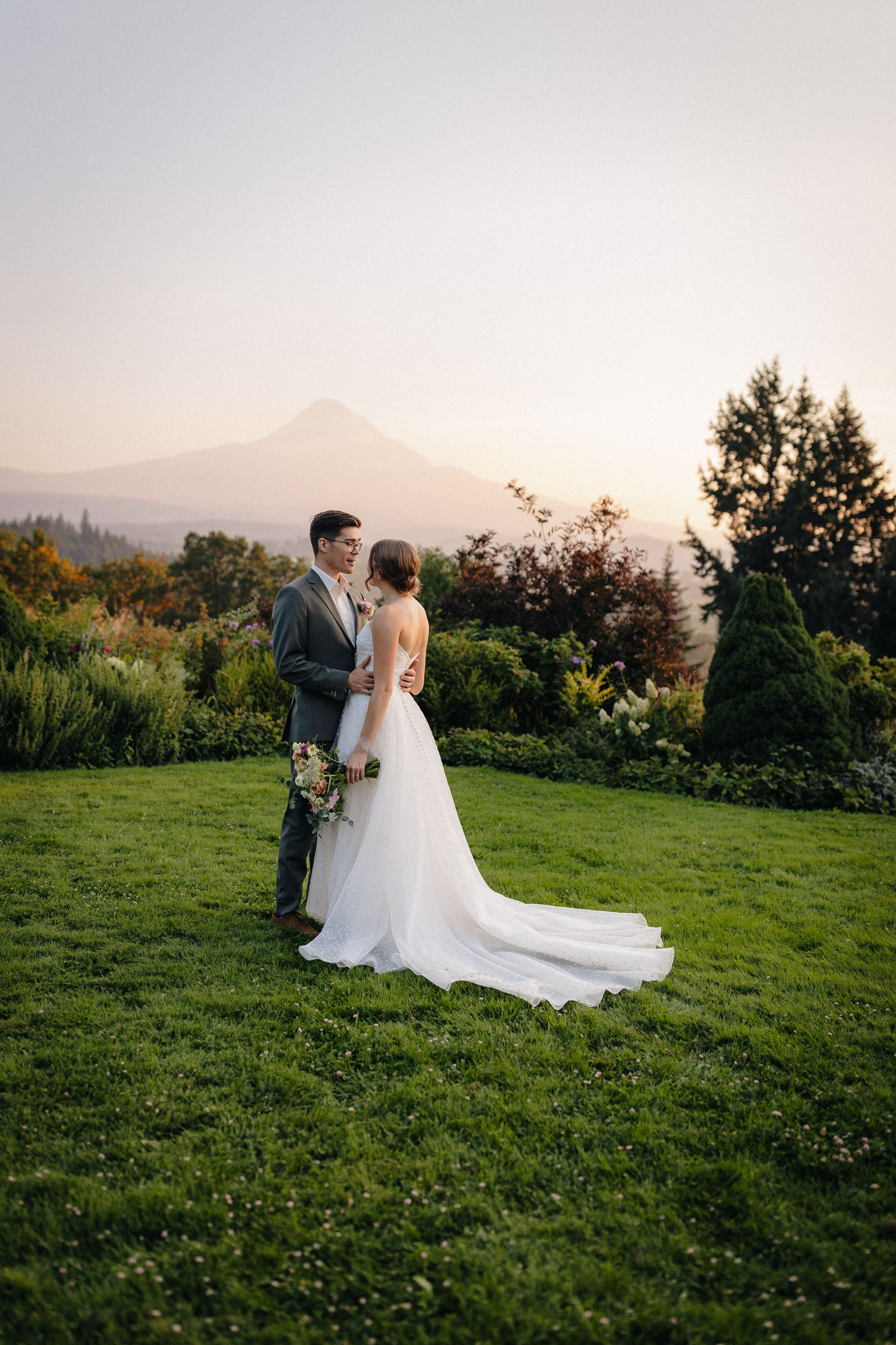 A bride and groom embrace on a hilltop lawn with mountain views and lush greenery at golden hour.
