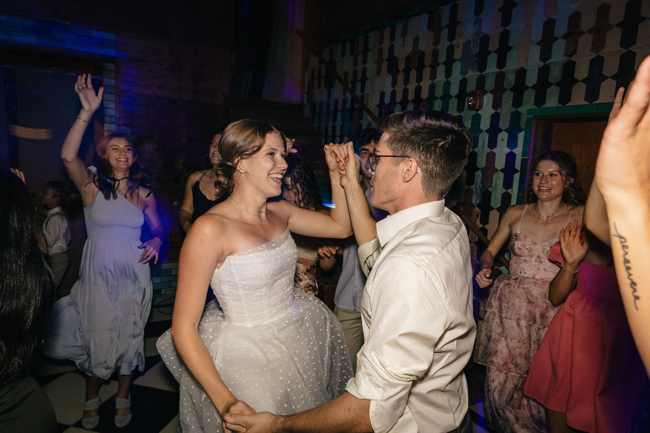 Two wedding guests dance together joyfully at a reception with blue lighting and other guests visible in background.