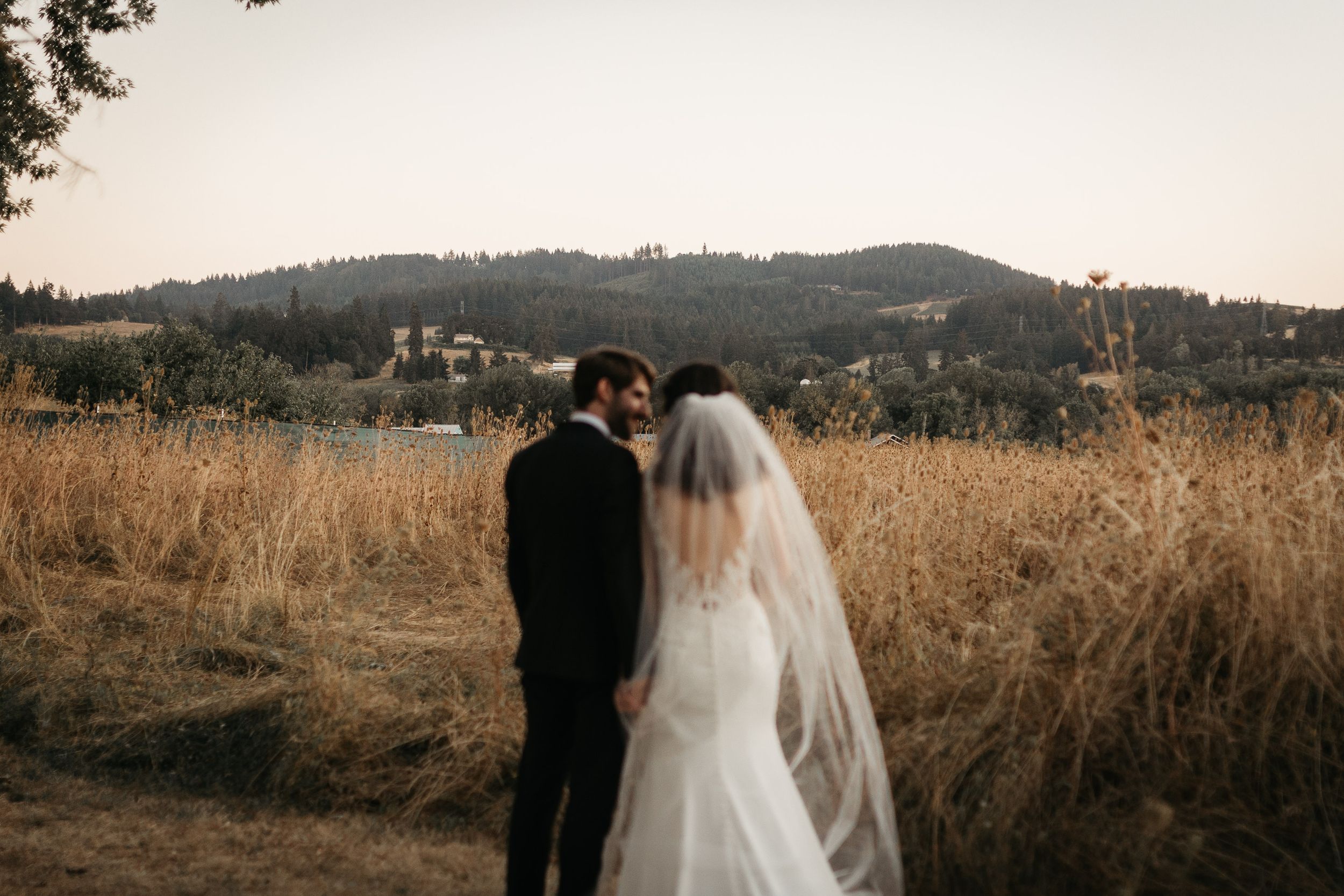 A couple in wedding attire stands together in a golden field with rolling hills and forest in the background at sunset.