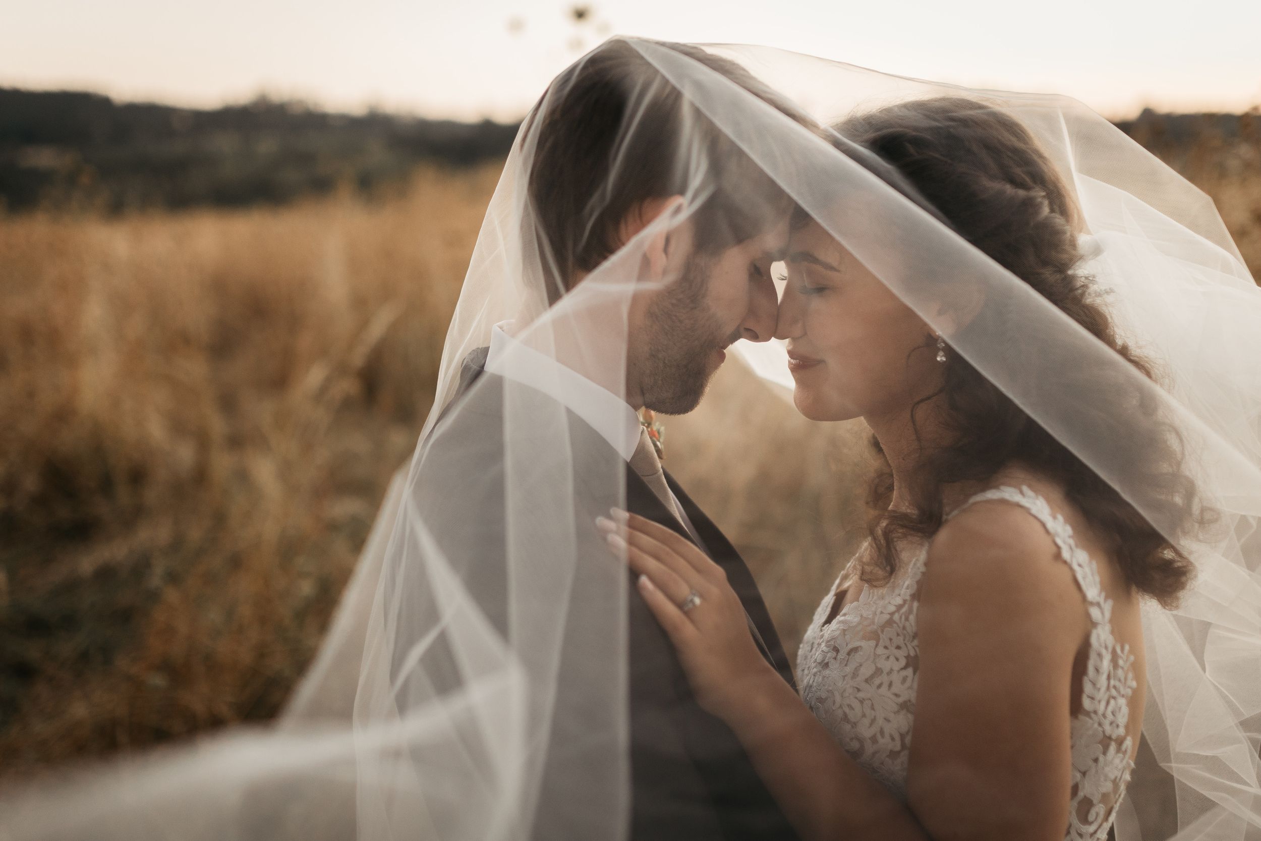 A romantic moment between newlyweds under a flowing veil during a golden hour photoshoot in a field.