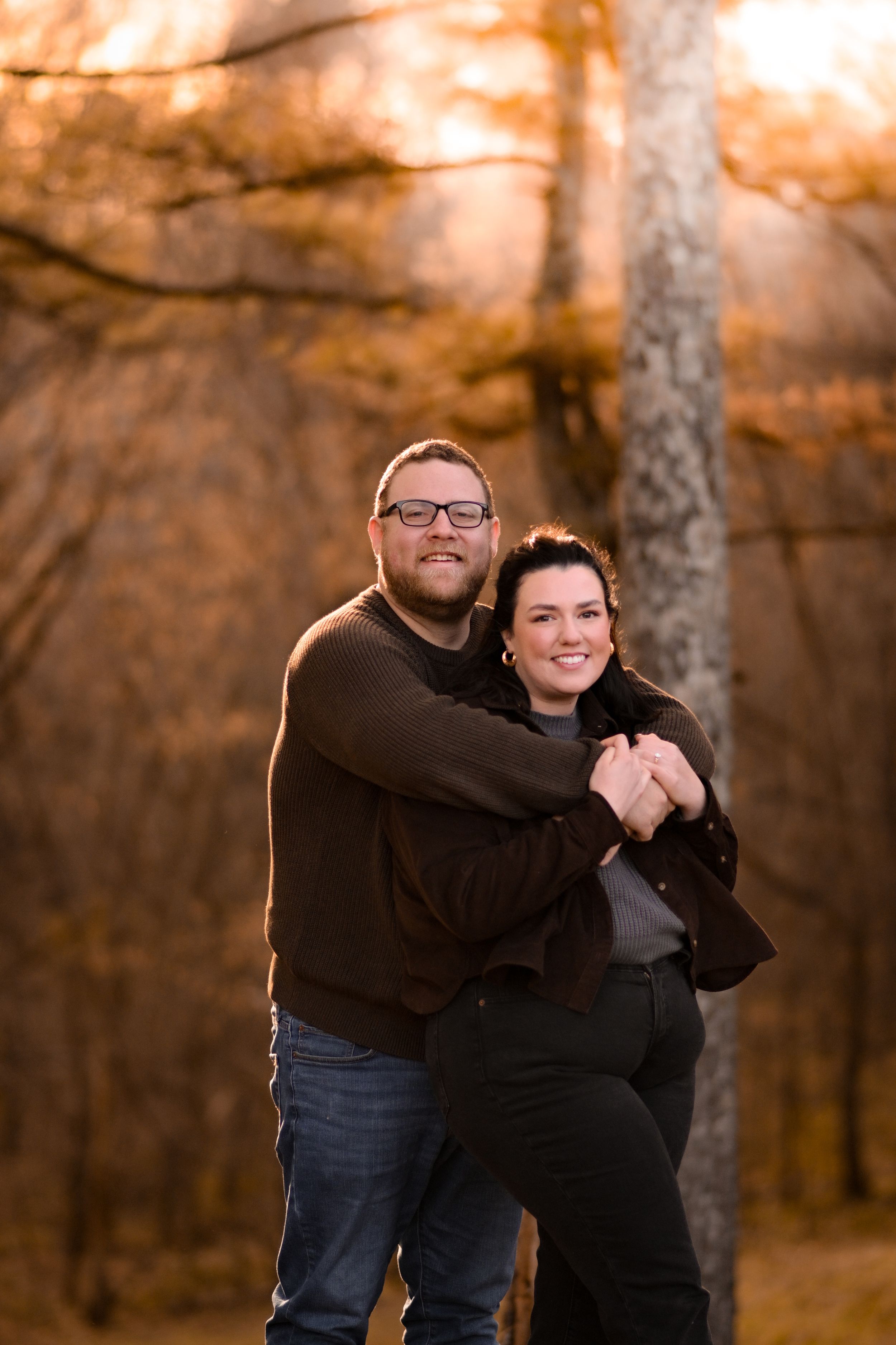 A couple embraces while standing among autumn trees at sunset.