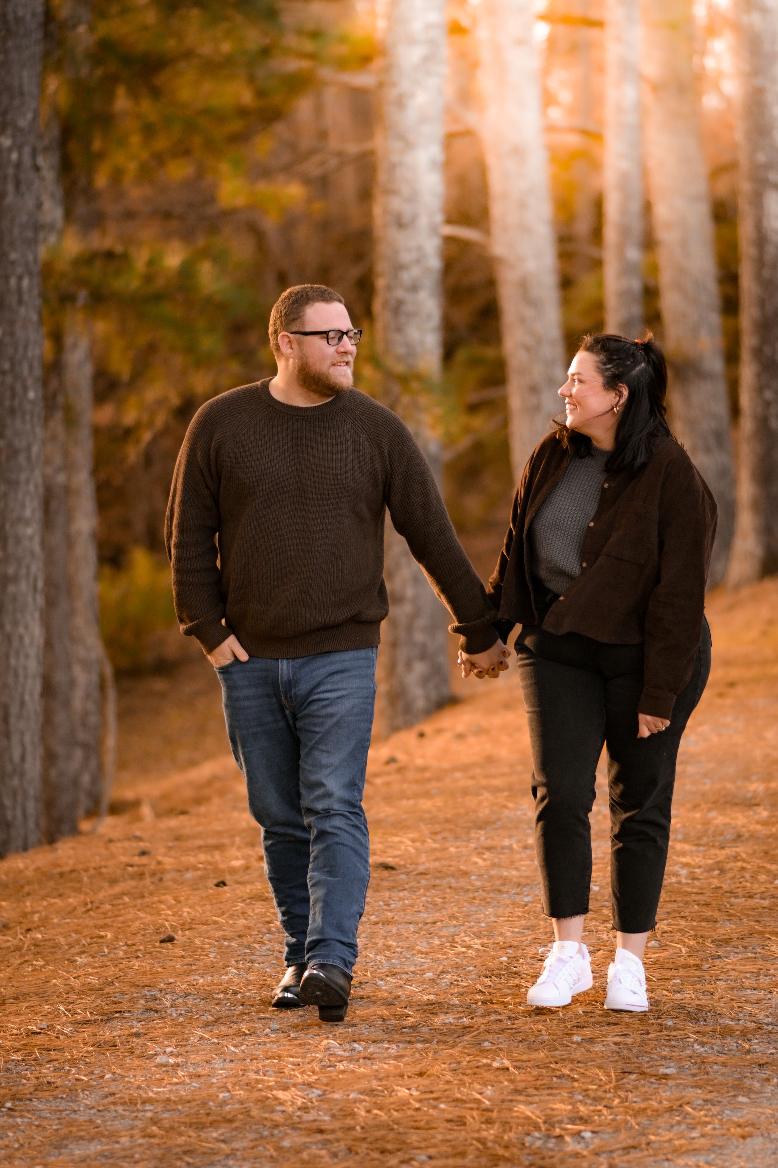 A couple walks hand in hand through a pine forest with golden sunlight filtering through.