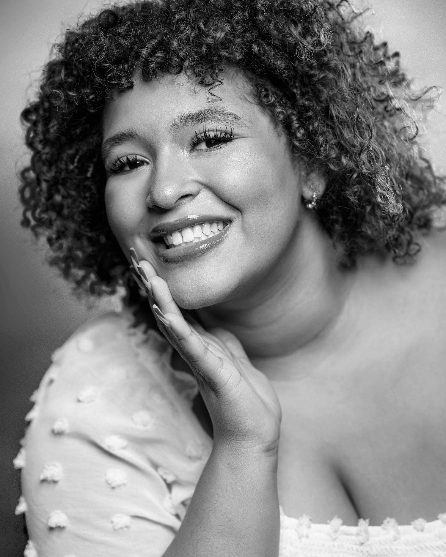 Black and white portrait of a smiling young woman with curly hair touching her cheek.