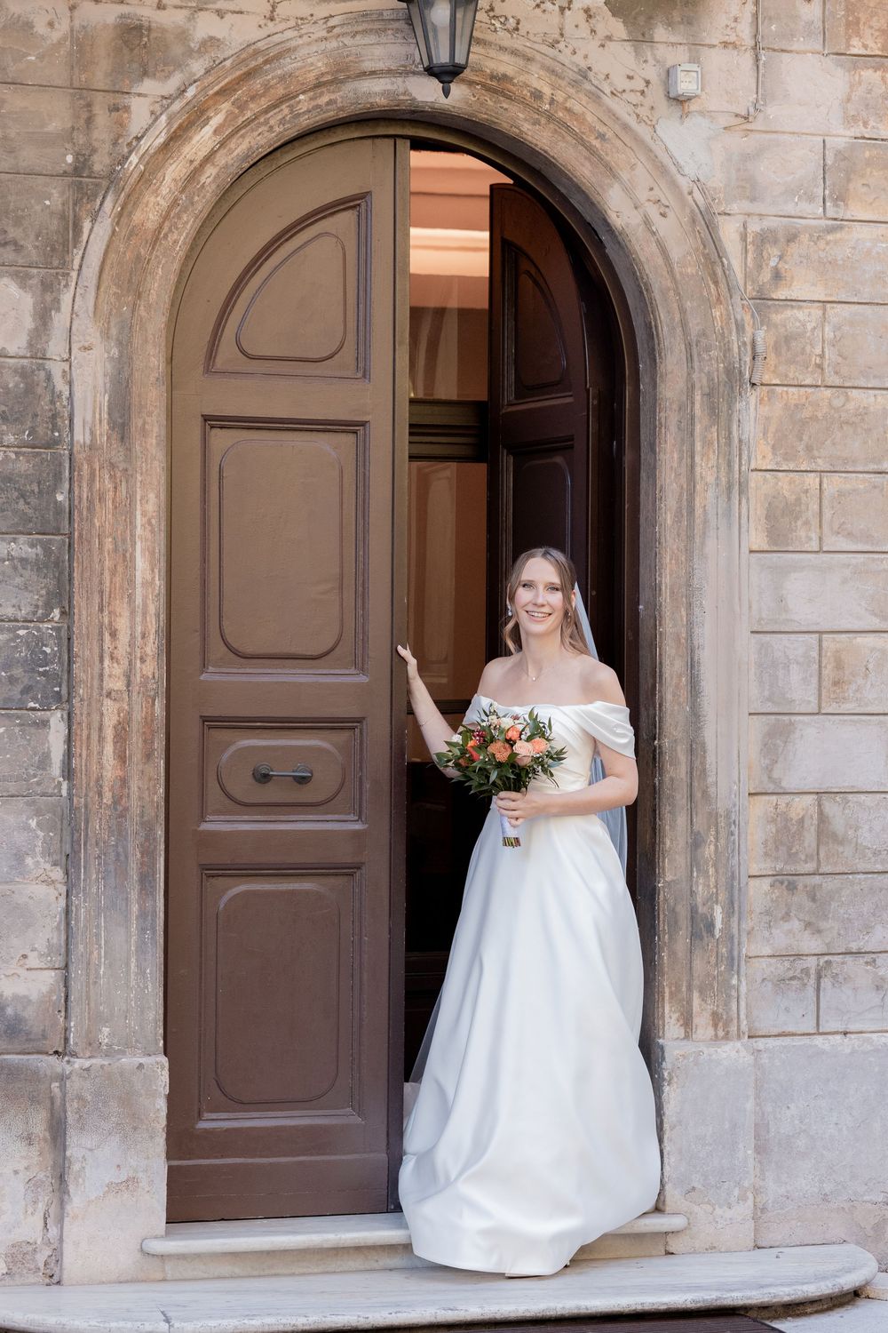 Smiling bride in an off-the-shoulder white wedding gown holding a bouquet while standing in an open arched wooden doorway of an old stone bu