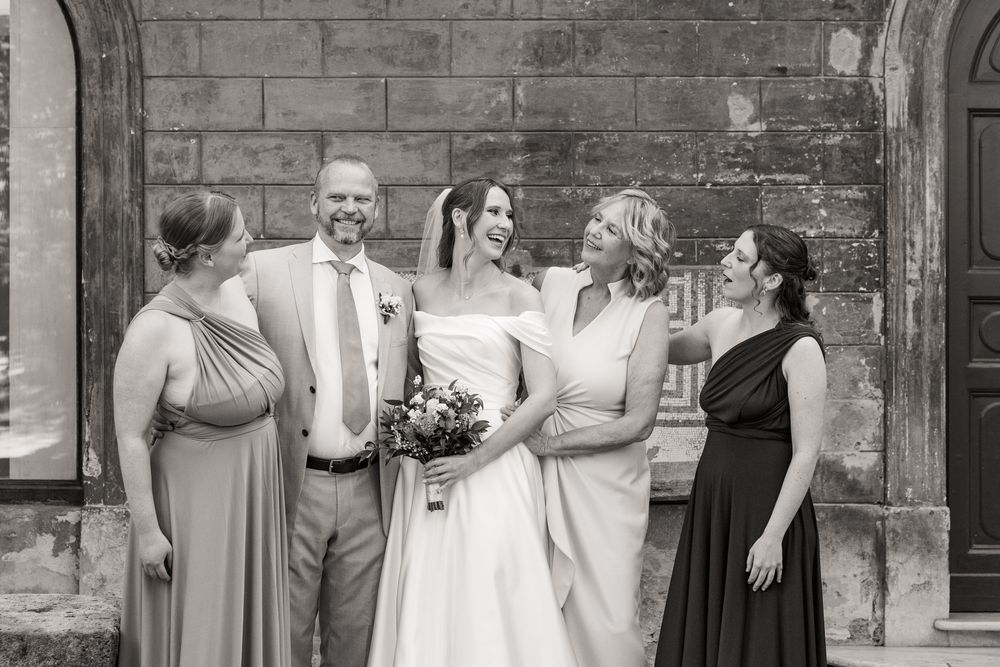 Smiling bride in a white gown holding a bouquet laughs with a groom and three bridesmaids in elegant dresses in front of a stone wall.