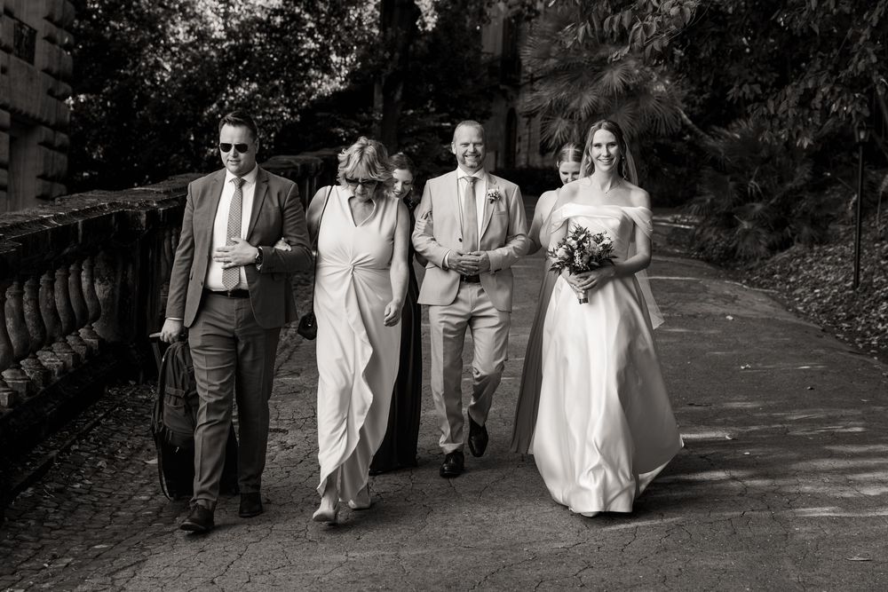 Black and white photo of a bride in a strapless gown holding a bouquet, walking down a garden path with family in formal wedding attire.