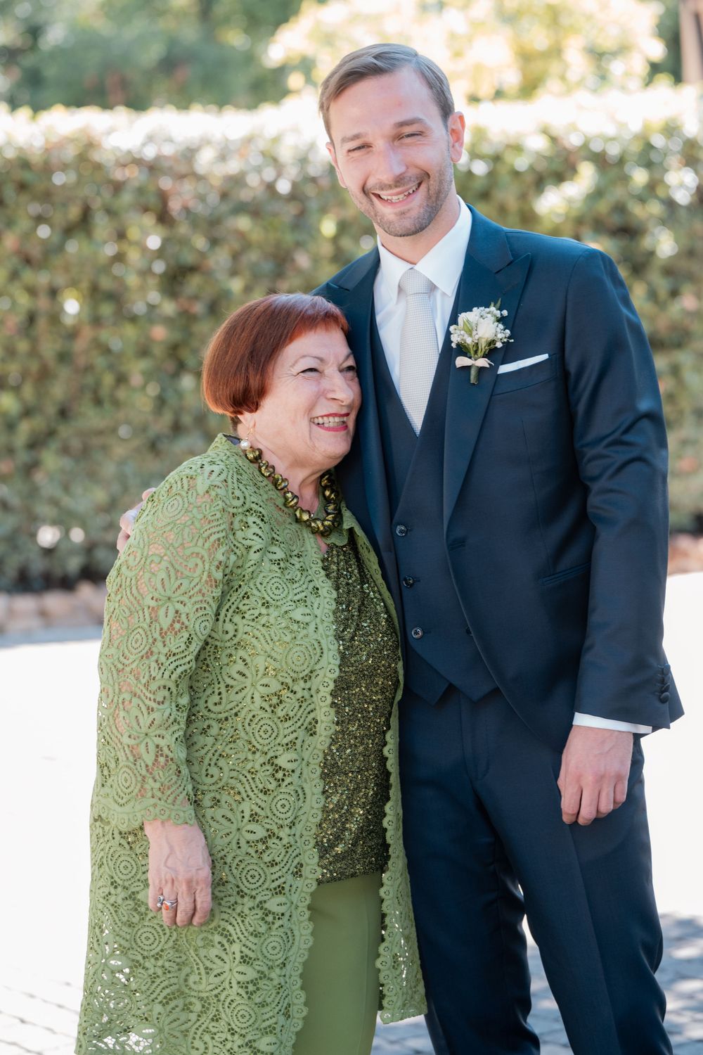 A smiling groom in a navy suit with a white boutonniere embraces an older woman in a green lace jacket outdoors at a wedding.
