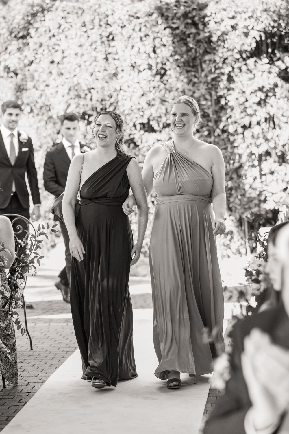 Two smiling bridesmaids in long one-shoulder dresses walk arm in arm down a wedding aisle past groomsmen in suits in black and white.