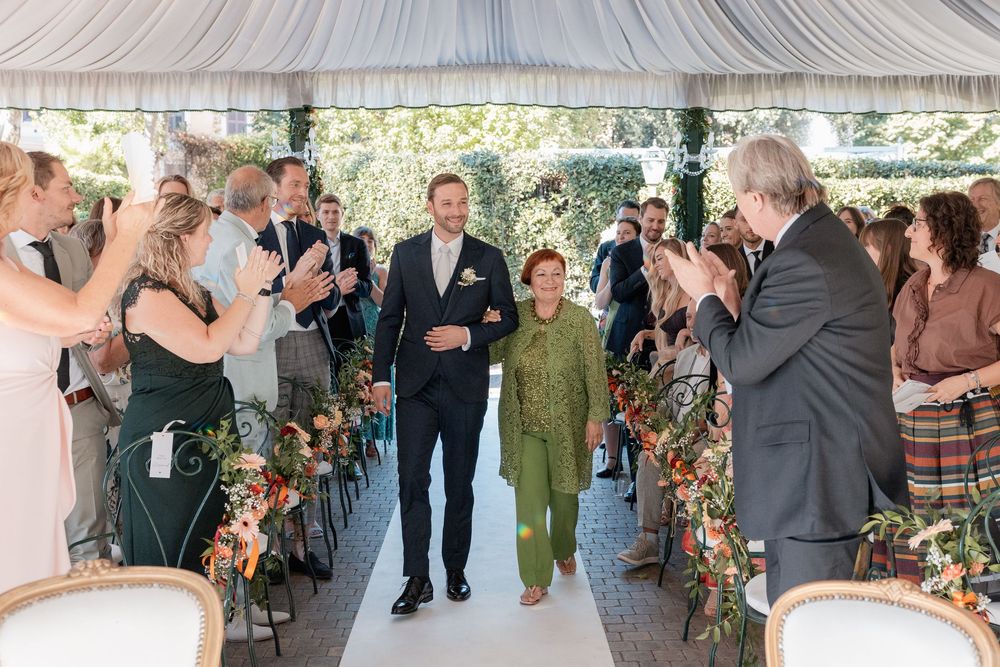 Groom in a dark suit walks an older woman in green down a flower-lined aisle as seated wedding guests applaud under a white tent.