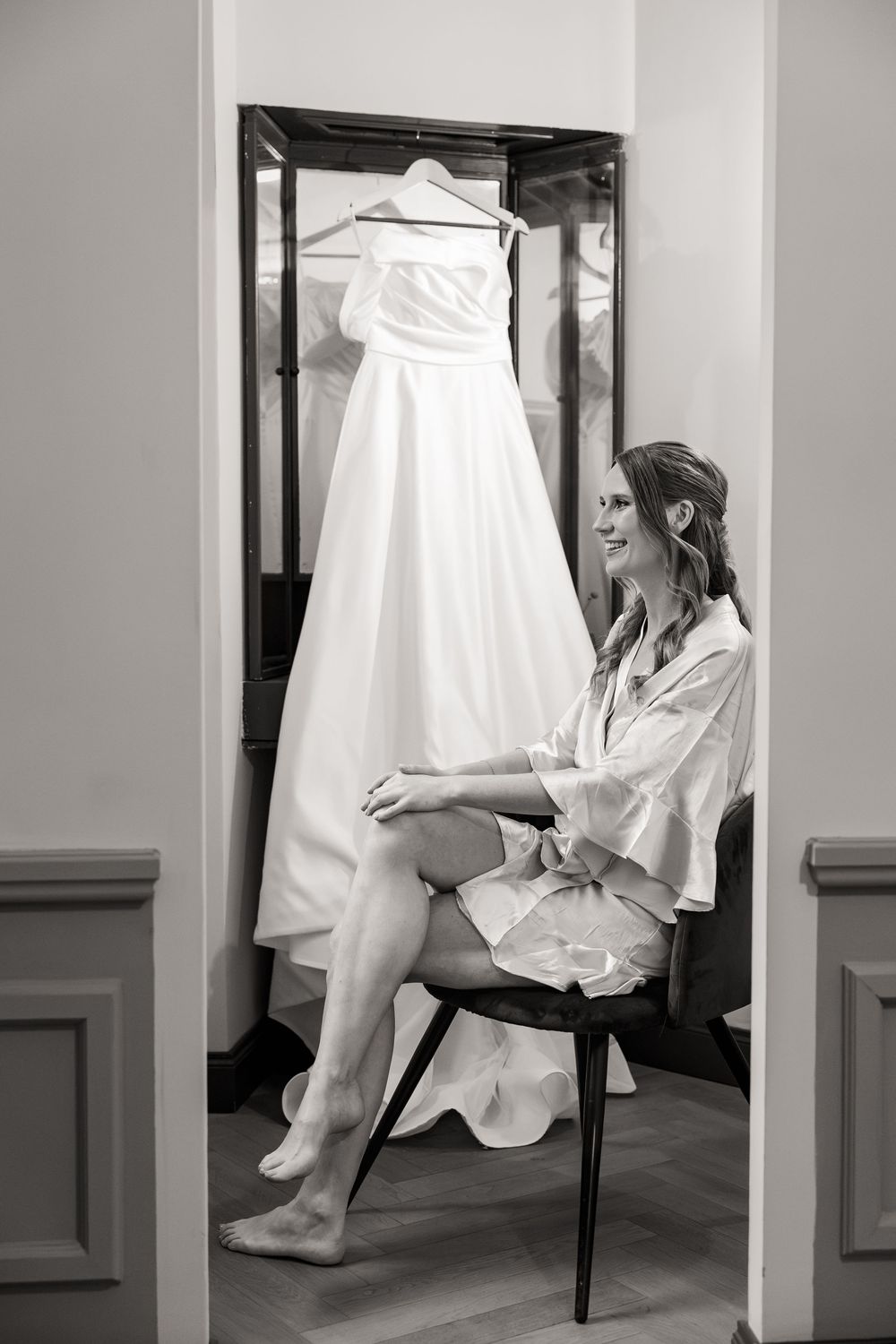 Smiling bride in a robe sits barefoot on a chair beside a white wedding gown hanging in a mirrored alcove, black and white photo.
