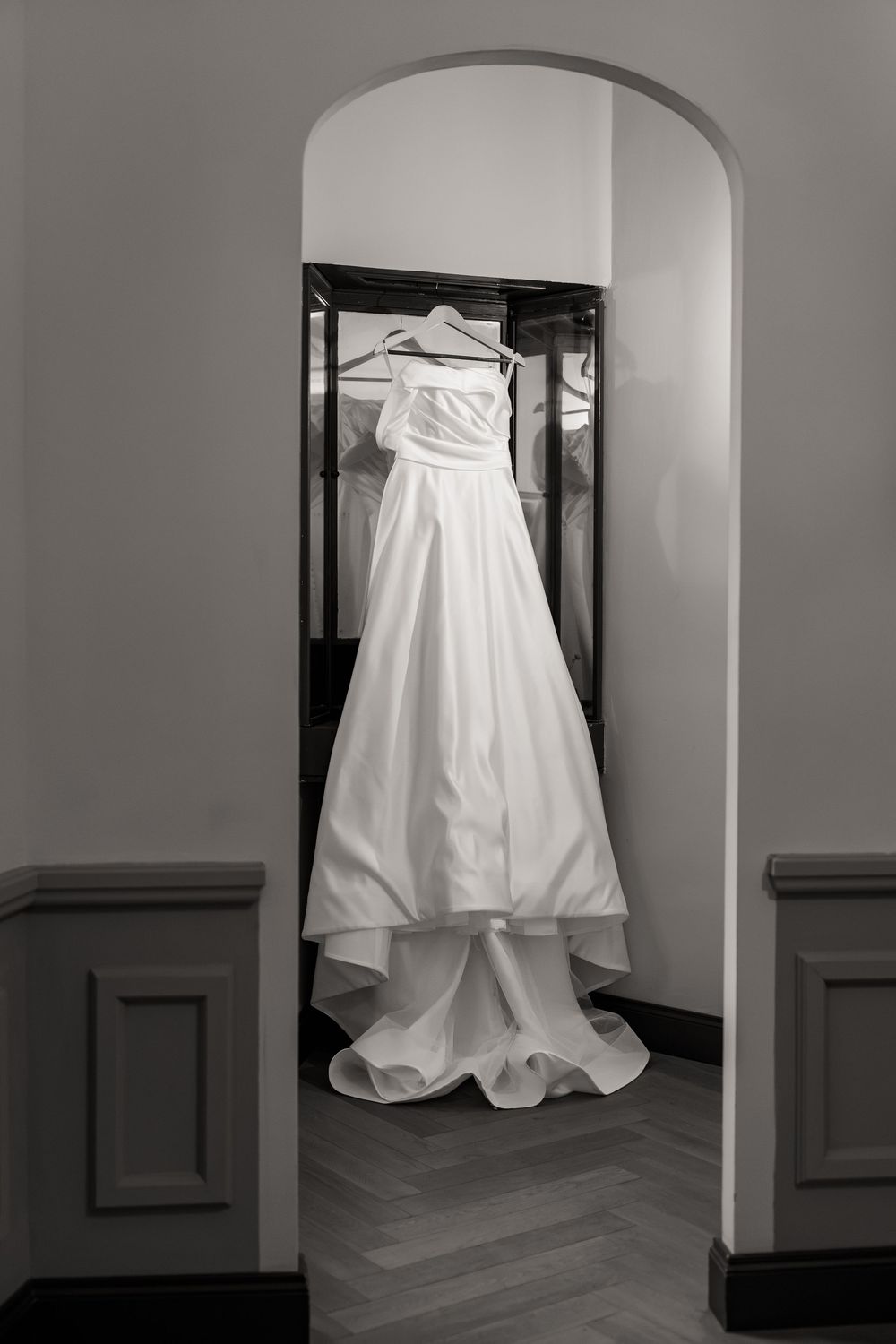 Strapless white wedding gown with a flowing train hangs on a hanger in an arched doorway in this black and white bridal photo.