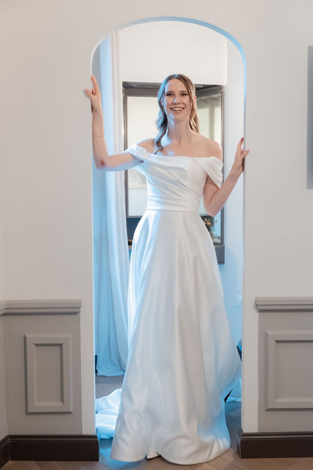 Smiling bride in an elegant off-the-shoulder satin wedding gown standing in an arched doorway with a flowing train.
