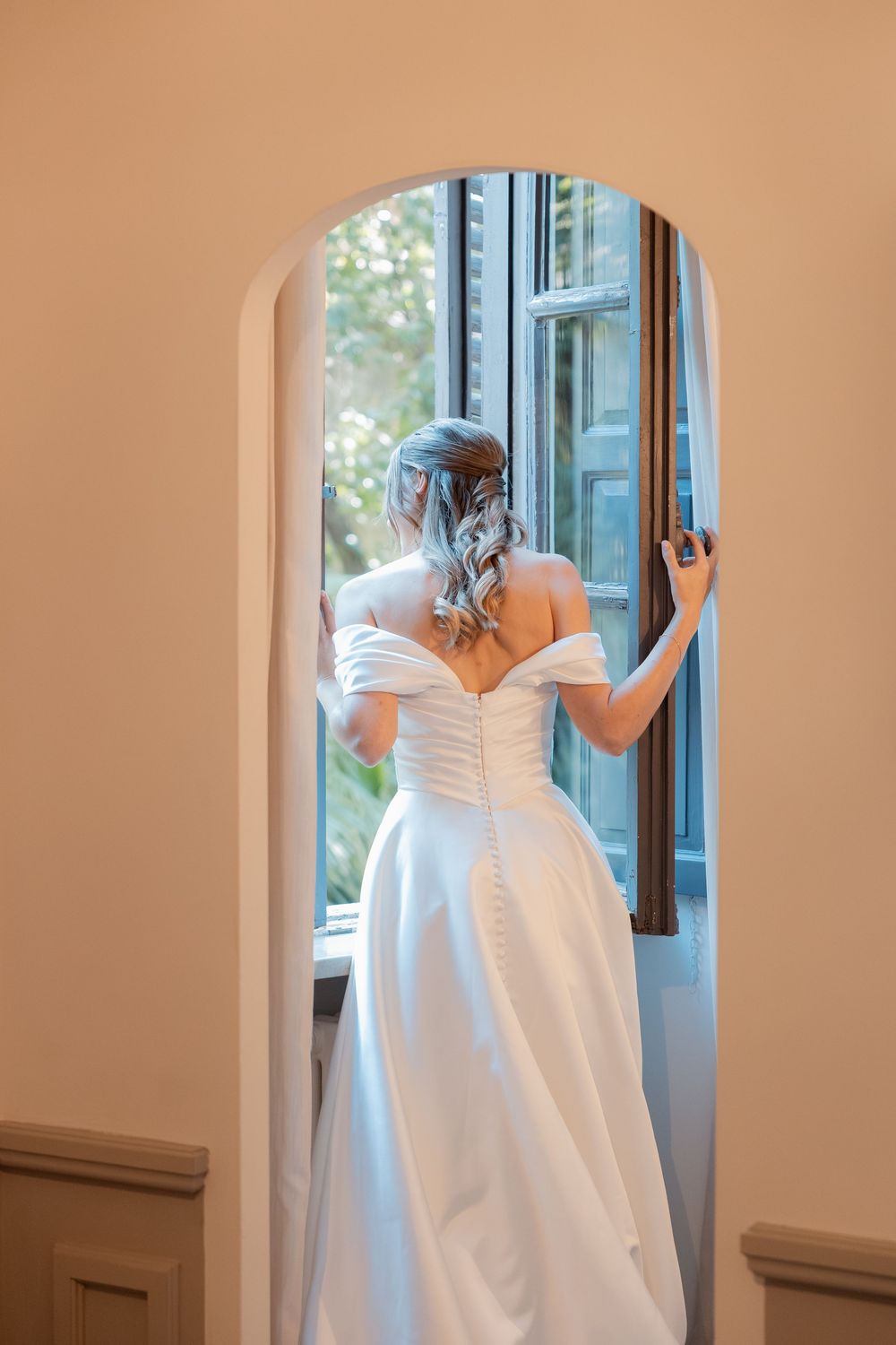Bride in an elegant off-the-shoulder satin wedding gown stands in an arched doorway, gazing out an open window with her hair in an updo.