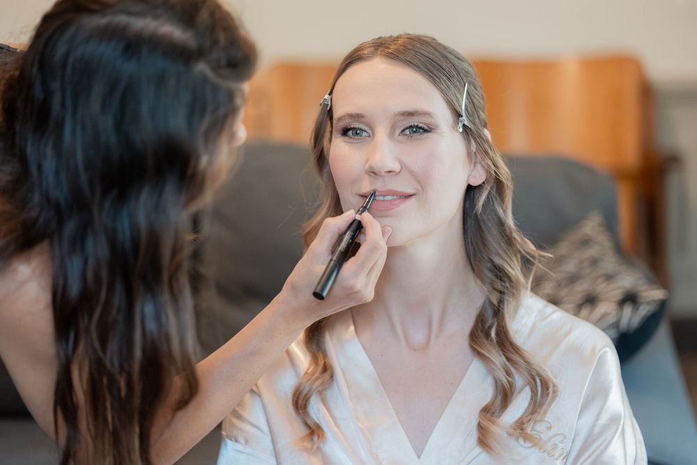 A makeup artist applies lip liner to a smiling woman with long wavy hair wearing a white satin robe during bridal makeup preparation.
