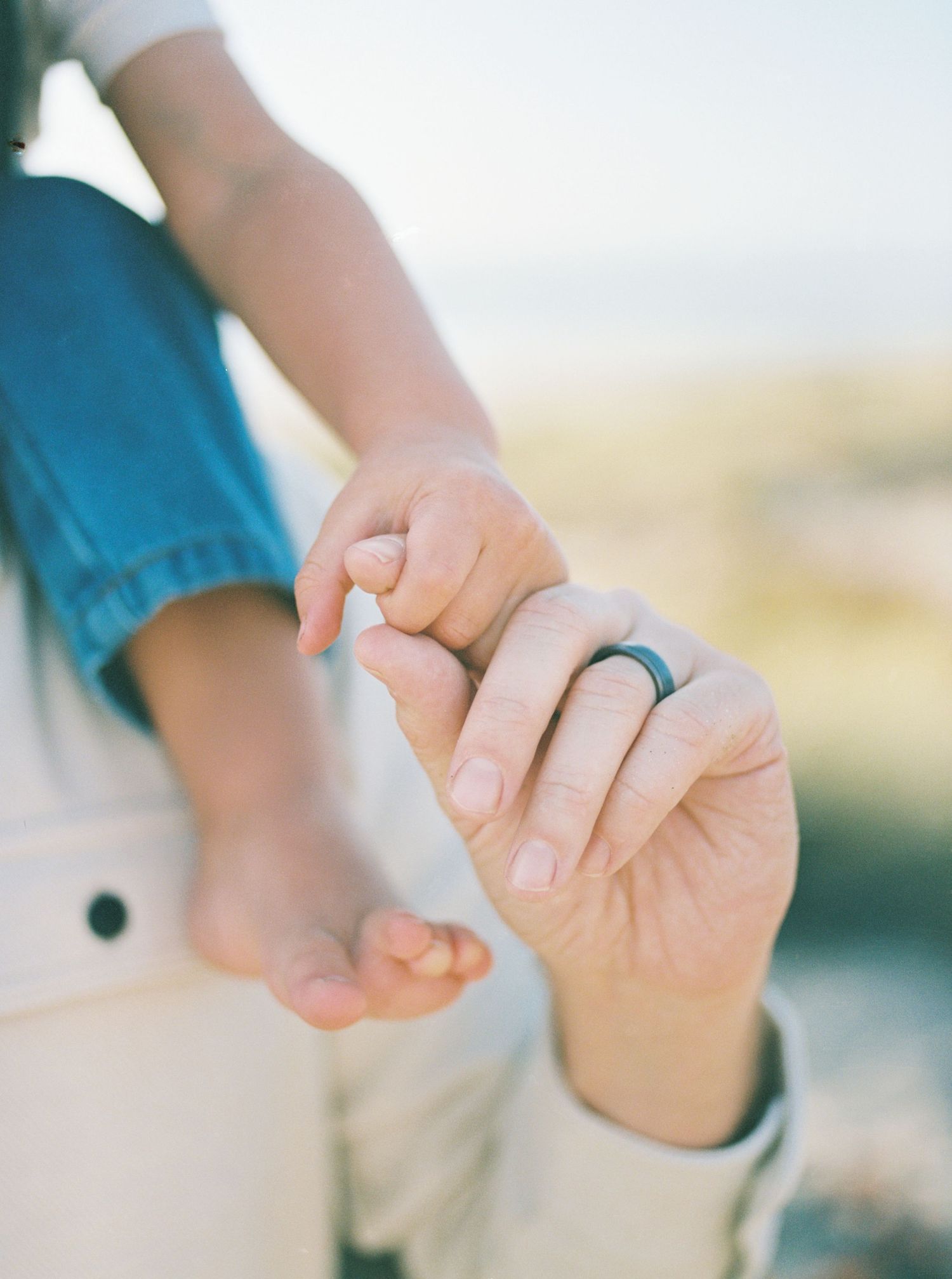 Adult hand wearing a dark ring gently holds a toddler's tiny hand and bare foot outdoors in soft, warm natural light.