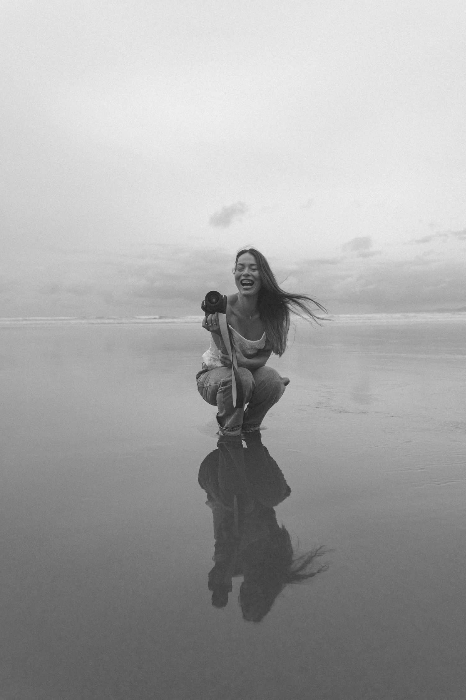 Black and white photo of a laughing woman crouching on a wet beach holding a camera, her hair blowing in the wind and reflected in the sand.
