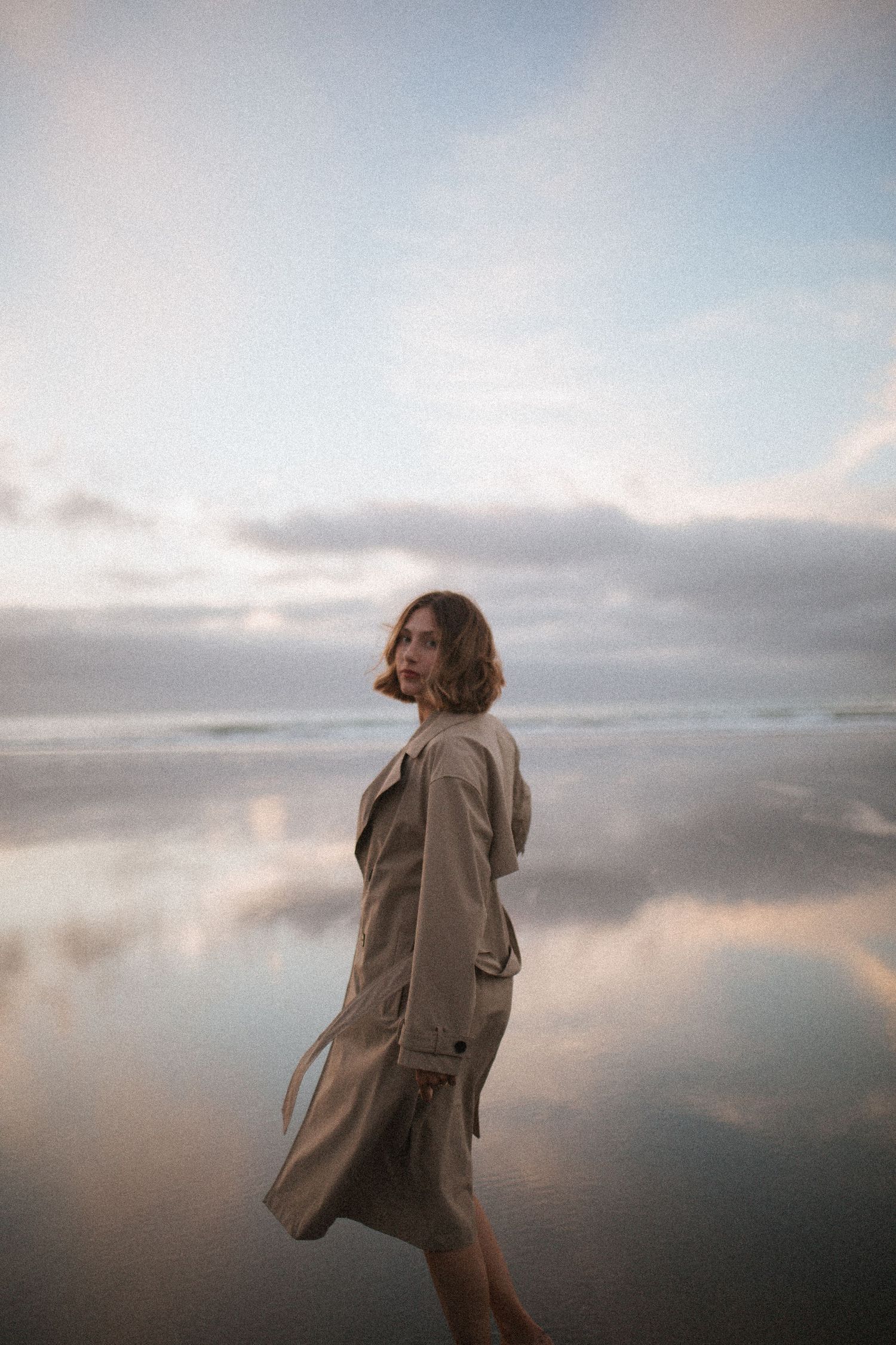 Woman in a tan trench coat looks over her shoulder on a wet beach, with soft pastel clouds reflected in the shallow water at dusk.