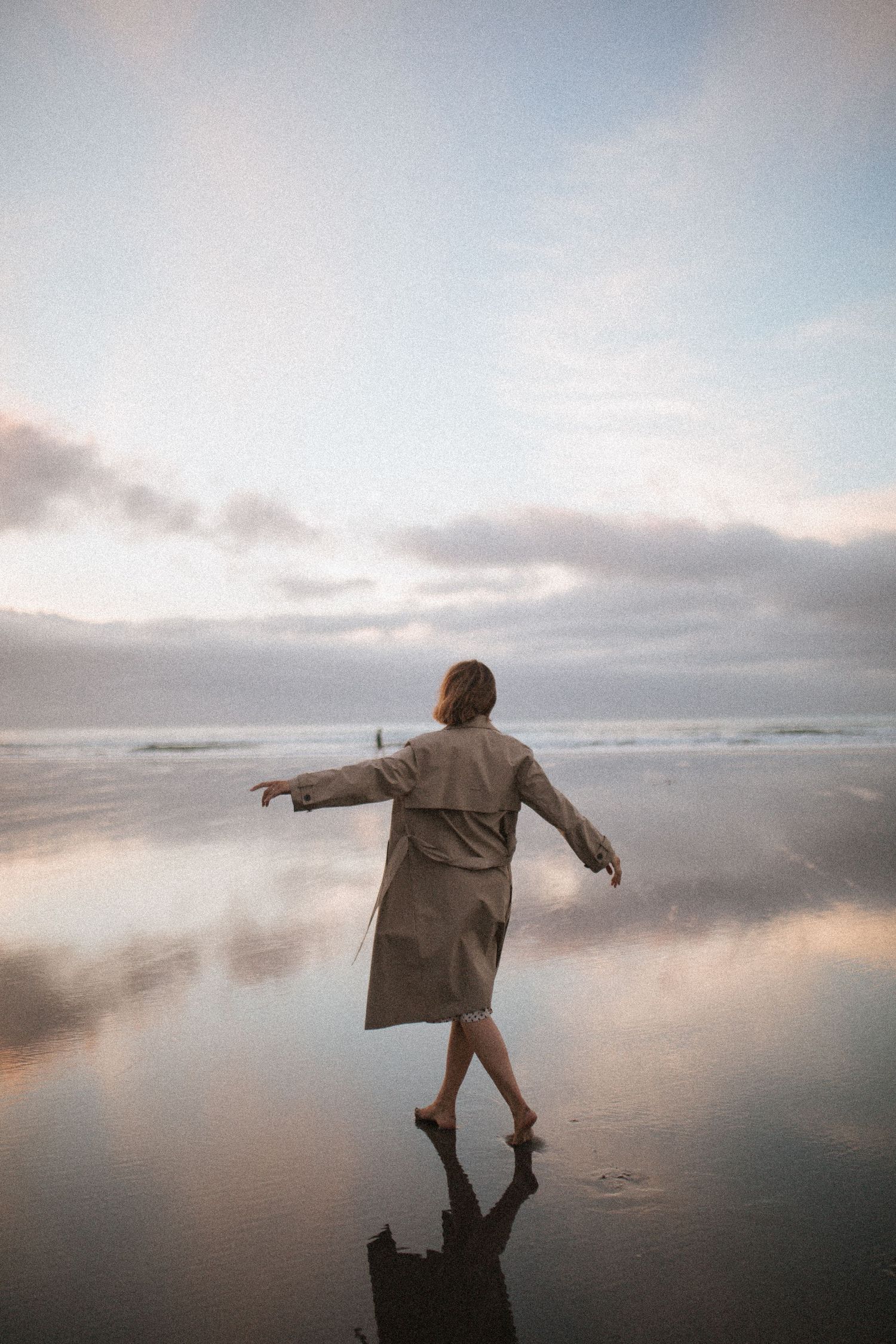 Woman in a beige trench coat walks barefoot with arms outstretched on a wet beach at sunset, reflected in the shimmering shallow water.
