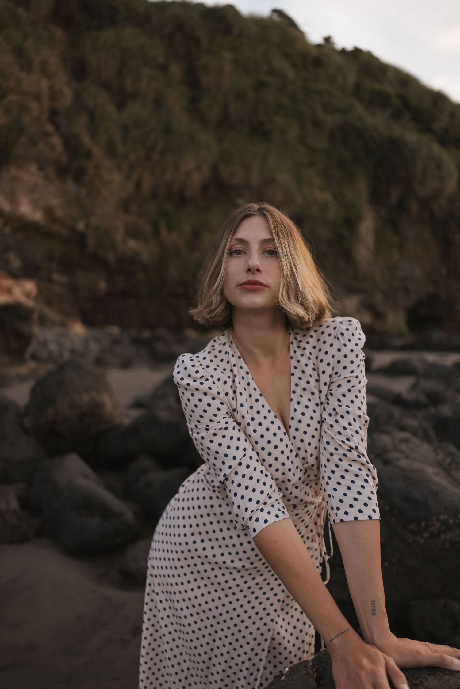 Woman with a short blonde bob wearing a cream polka dot wrap dress poses among dark coastal rocks with a green hillside behind her.