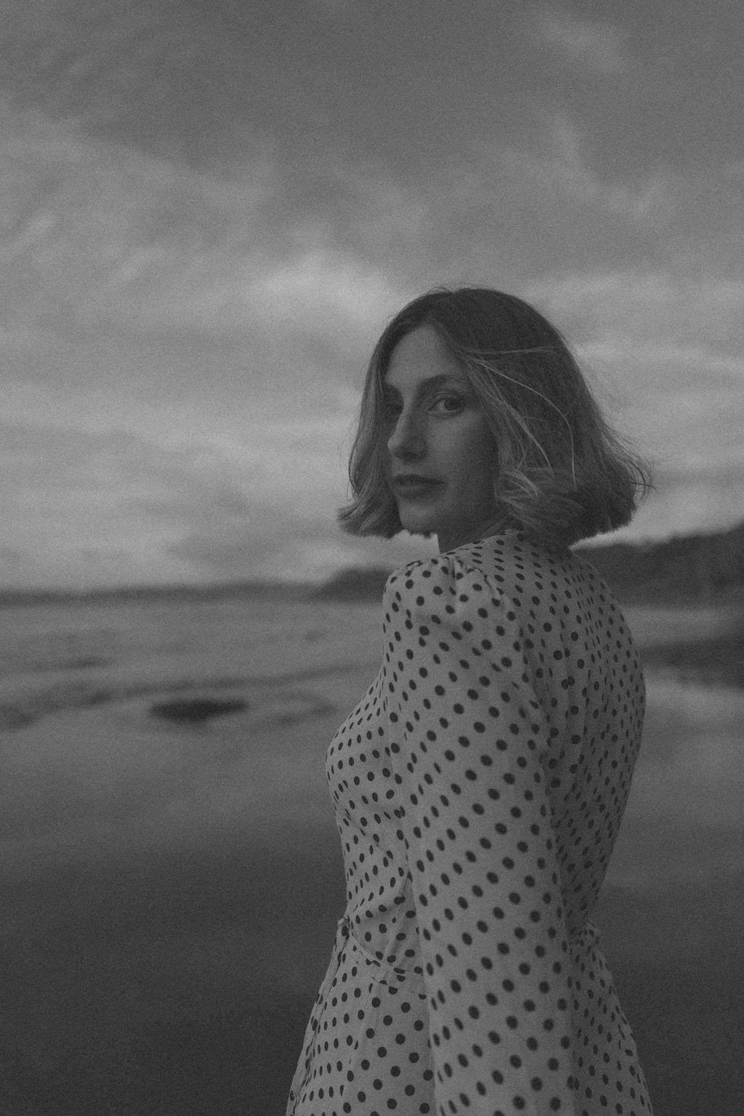 Black and white portrait of a woman with a wavy bob in a polka dot dress looking over her shoulder on a moody beach under cloudy skies.